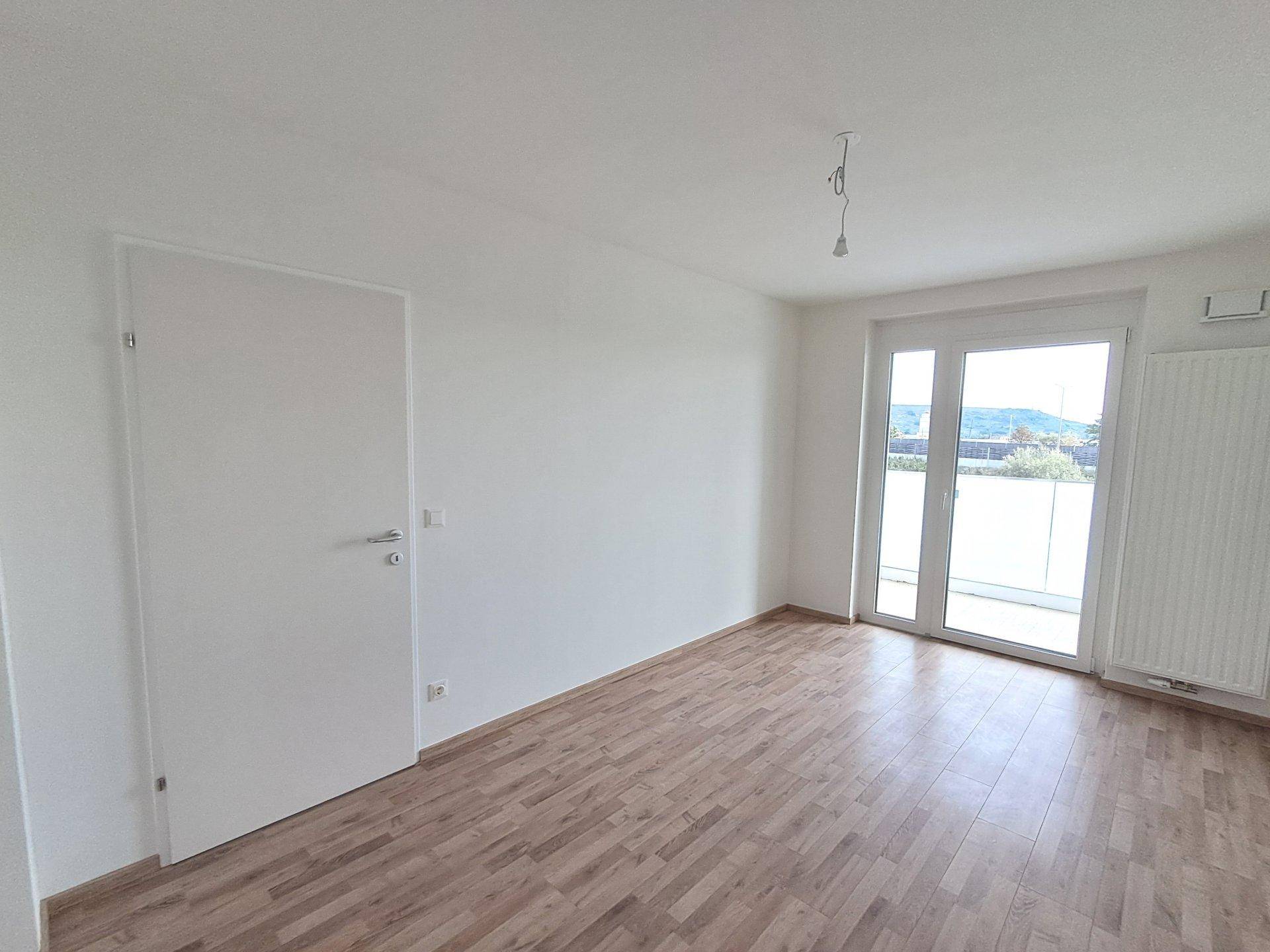 Heller, leerer Raum mit Holzboden und großen Fenstern, die Zugang zu einem Balkon bieten und den Raum erhellen.