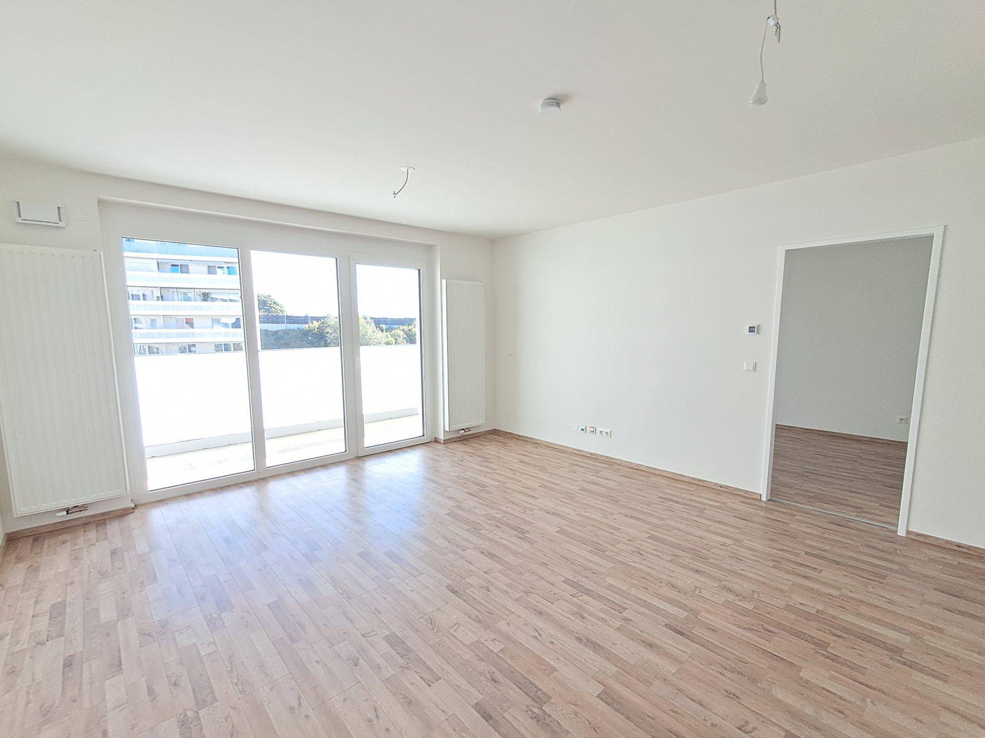 Heller, leerer Raum mit Holzboden und großen Fenstern, die viel Tageslicht hereinlassen und einen Ausblick bieten.
