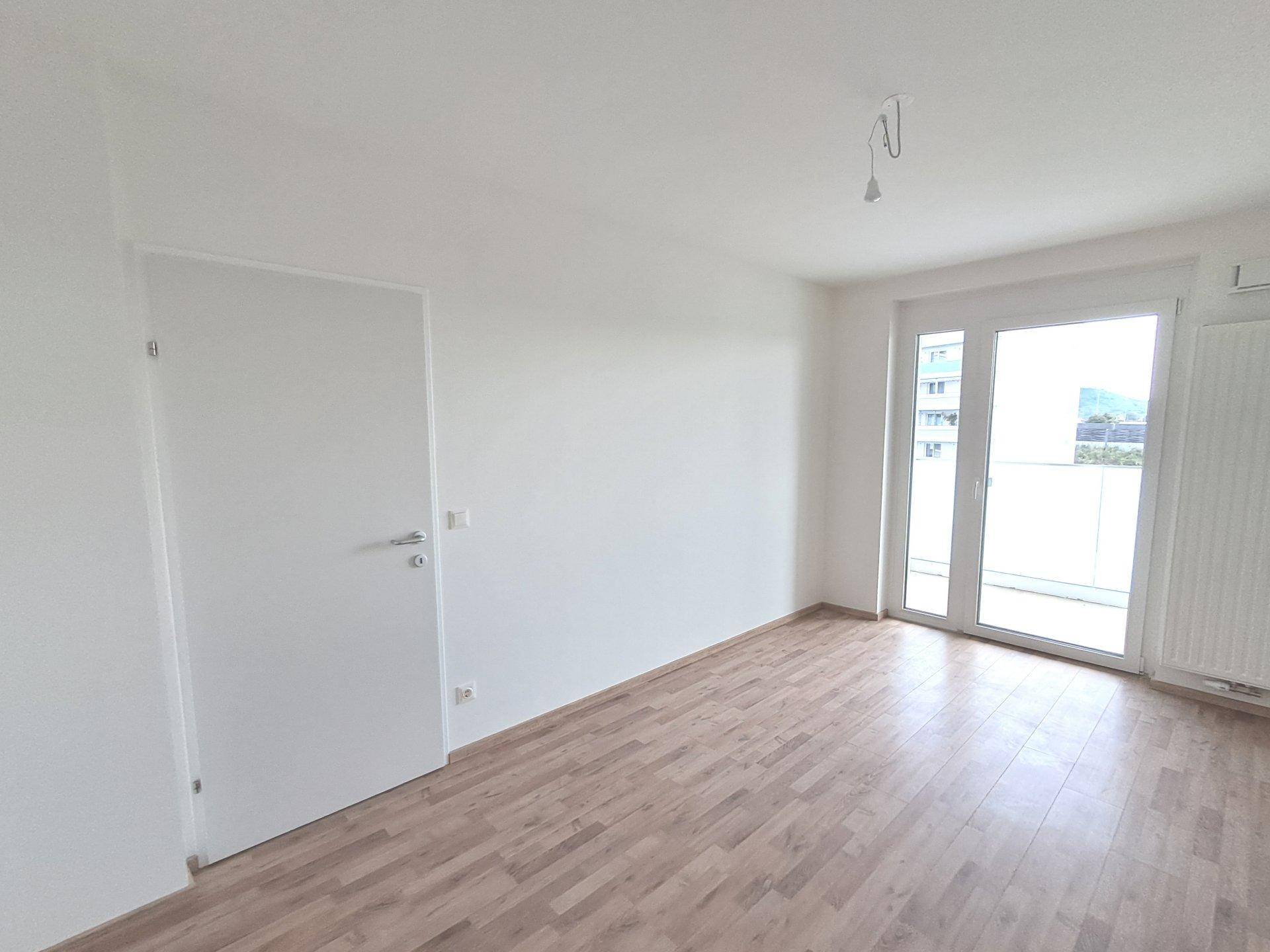 Leeres Schlafzimmer mit Laminatboden und Fensterfront, die Zugang zum Balkon bietet.