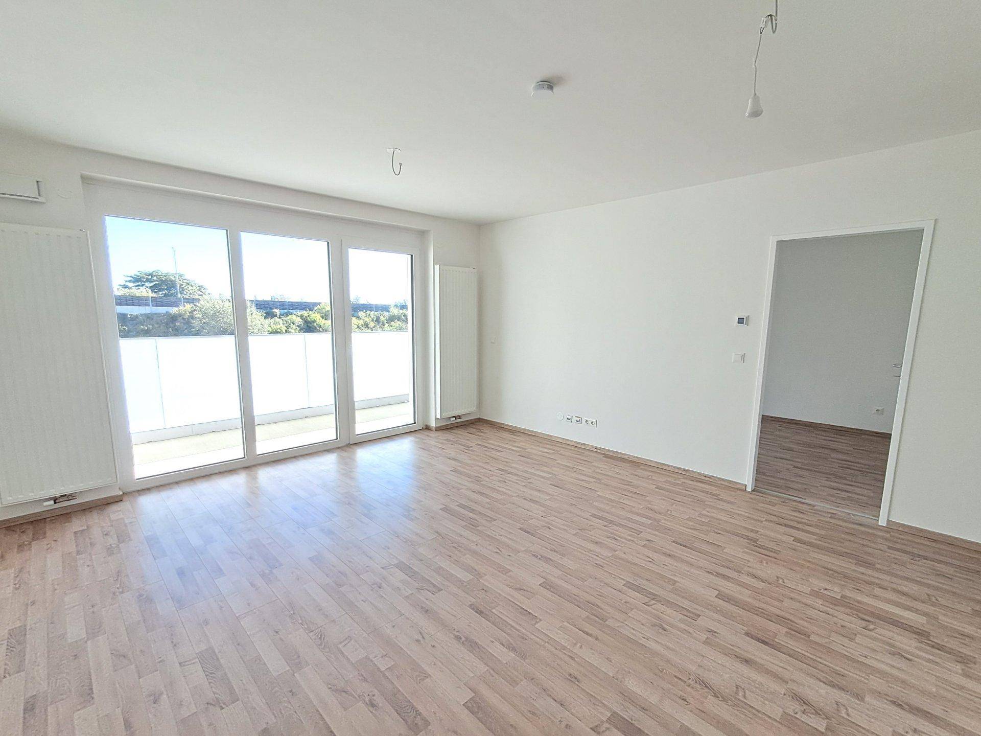 Geräumiges, leeres Wohnzimmer mit Holzboden und breiter Fensterfront für viel Helligkeit und Balkonzugang.