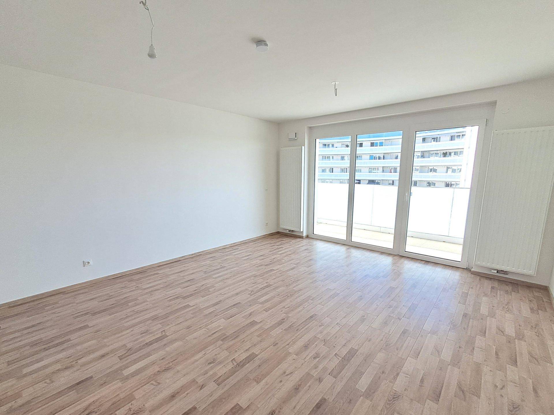 Helles, leeres Wohnzimmer mit Holzboden und großen Fenstern, die viel Tageslicht hereinlassen.