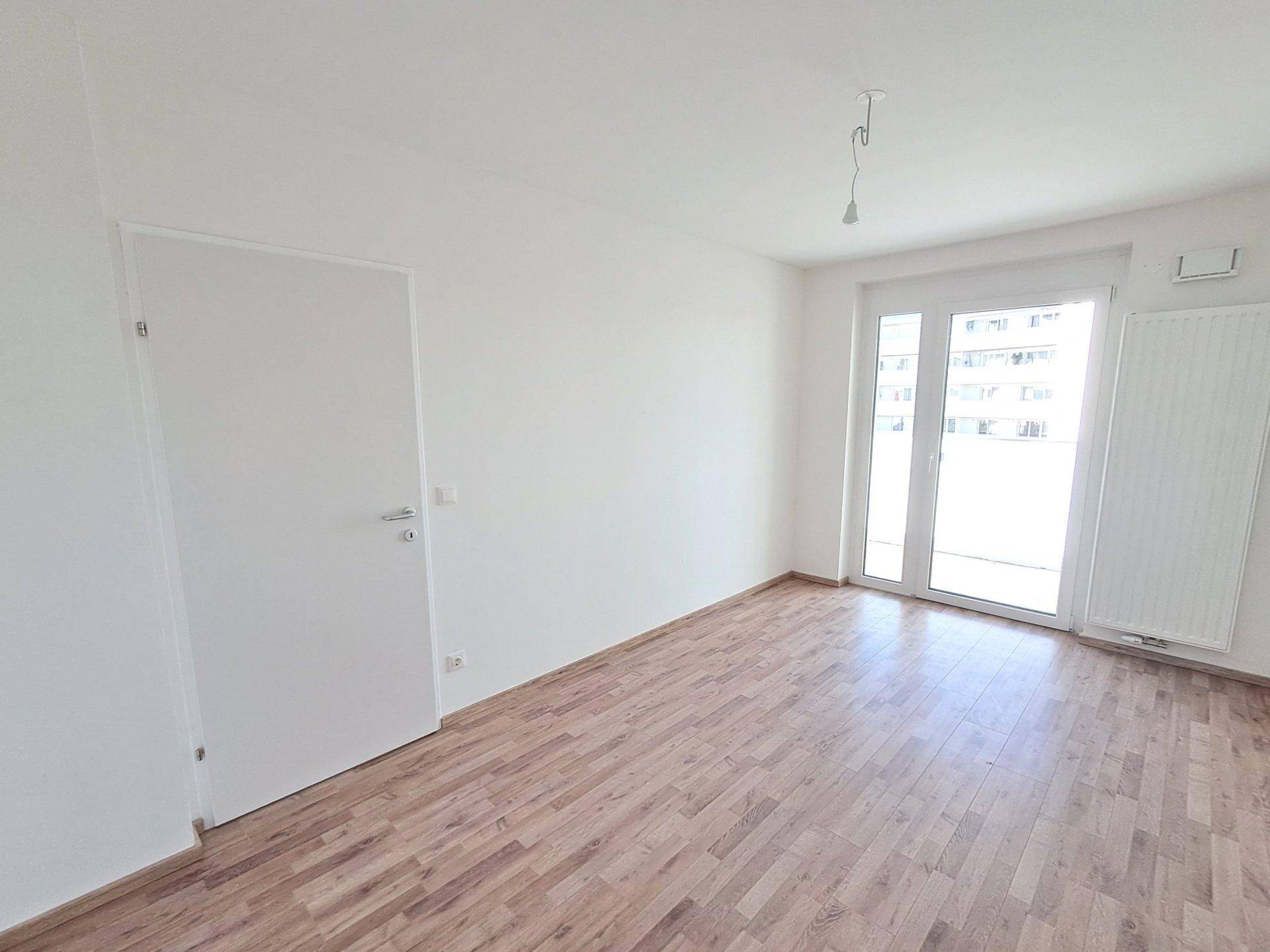 Helles, leeres Schlafzimmer mit Holzboden und Balkonzugang durch große Fenster.