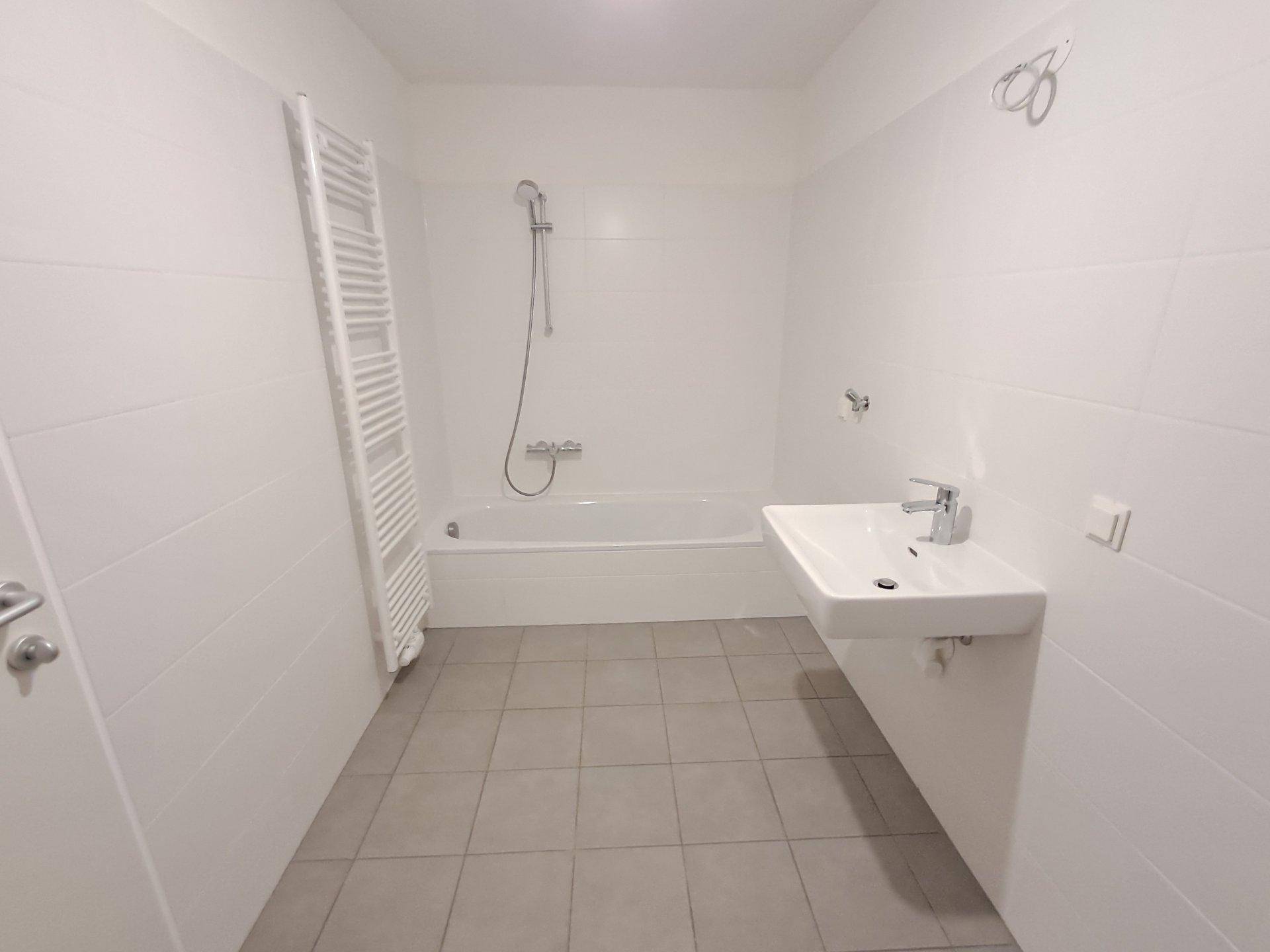 Helles Badezimmer mit weißen Fliesen, Badewanne mit Duschfunktion und beheiztem Handtuchhalter.