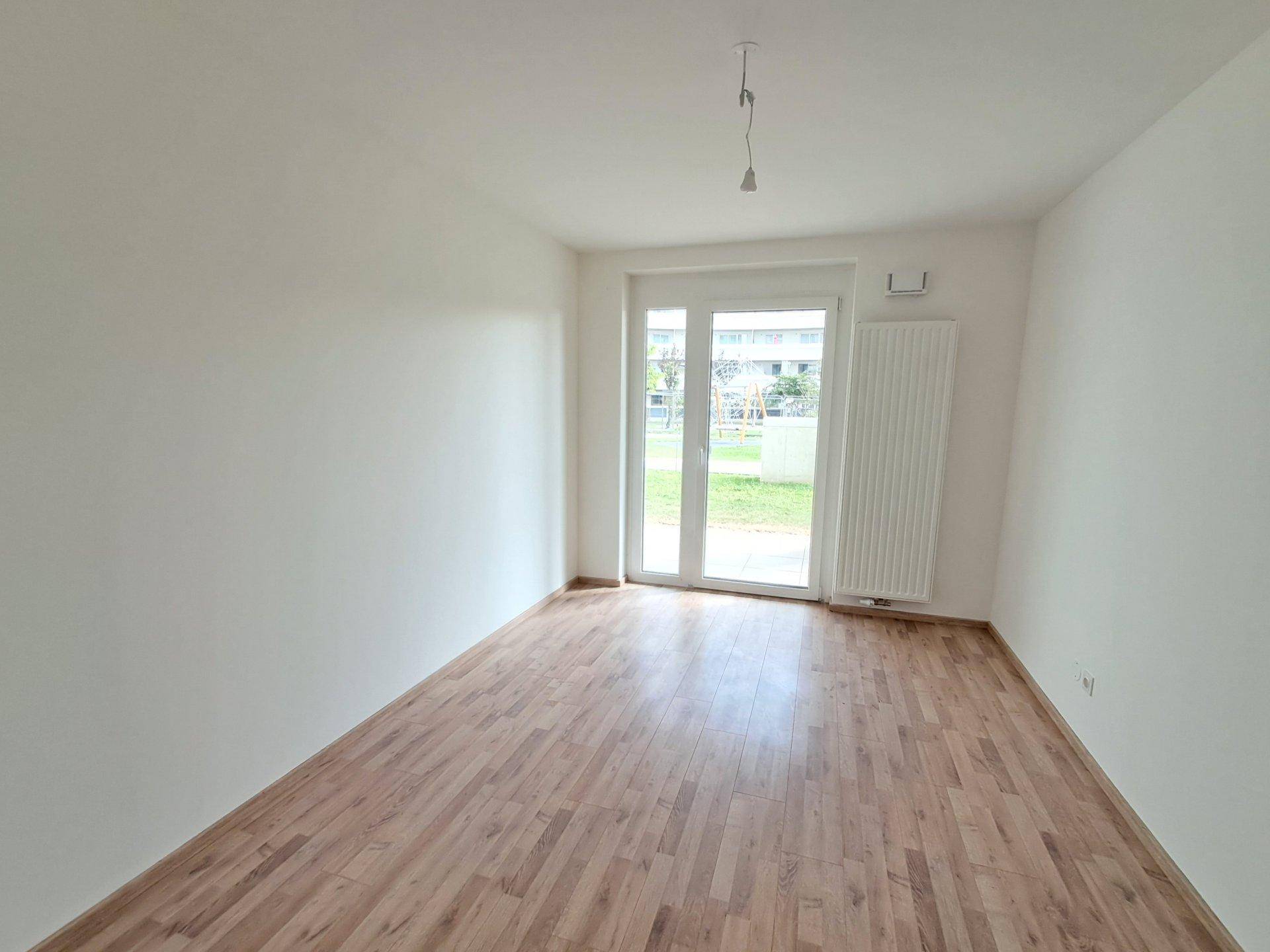 Leeres Zimmer mit Laminatboden und großem Fenster, das Zugang zu einem Außenbereich bietet.