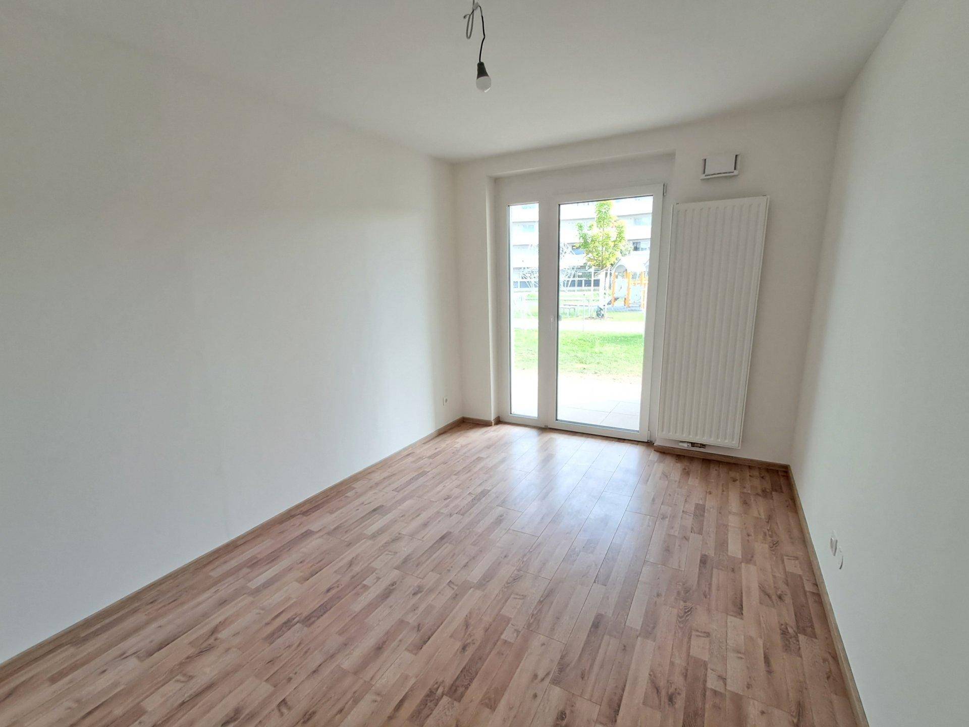Leeres Zimmer mit Laminatboden und großem Fenster, das viel Tageslicht hereinlässt und einen Blick ins Grüne bietet.