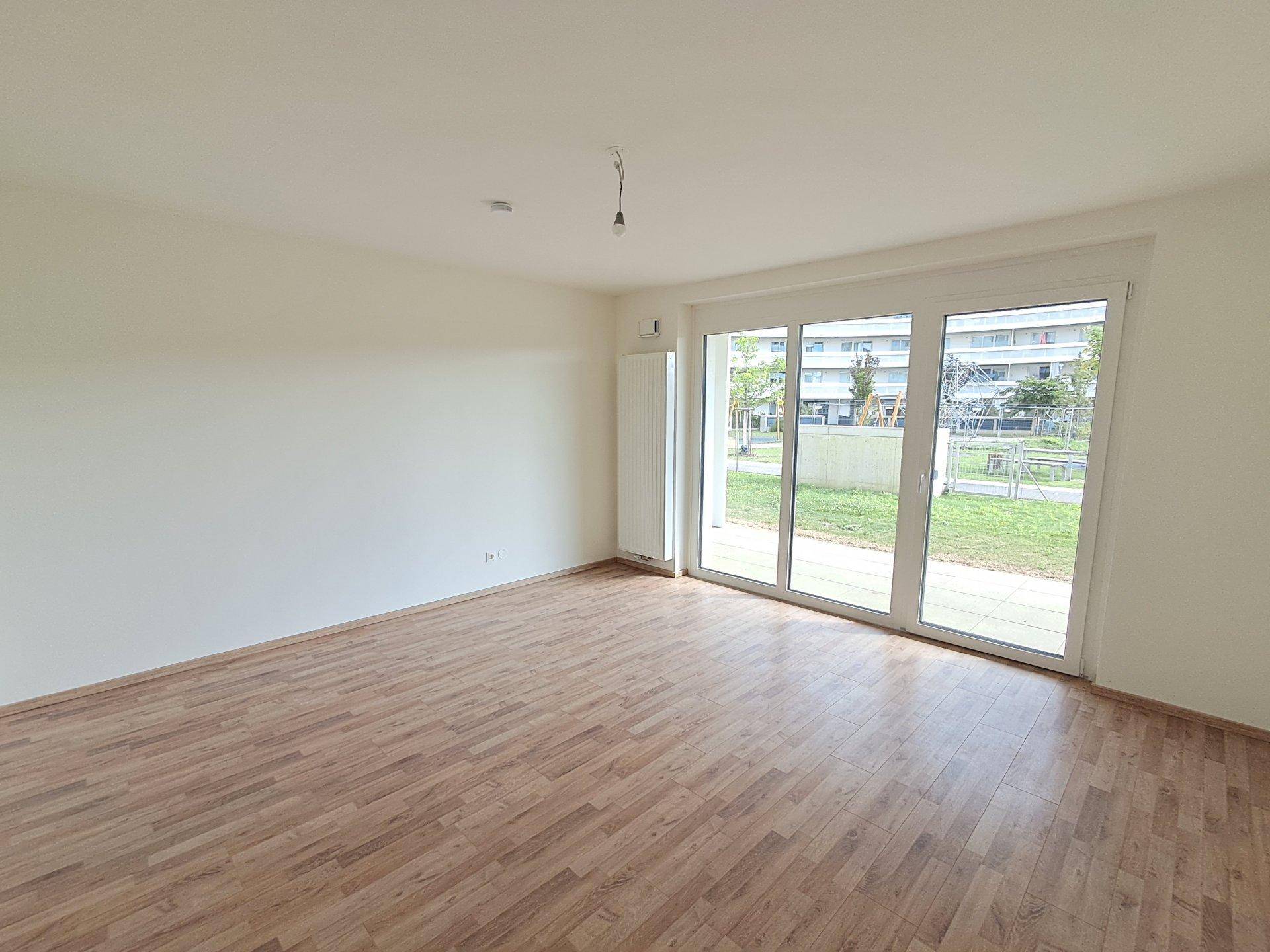 Helles Zimmer mit Laminatboden und großen Fenstern, die einen Blick auf die Grünfläche und andere Gebäude bieten.
