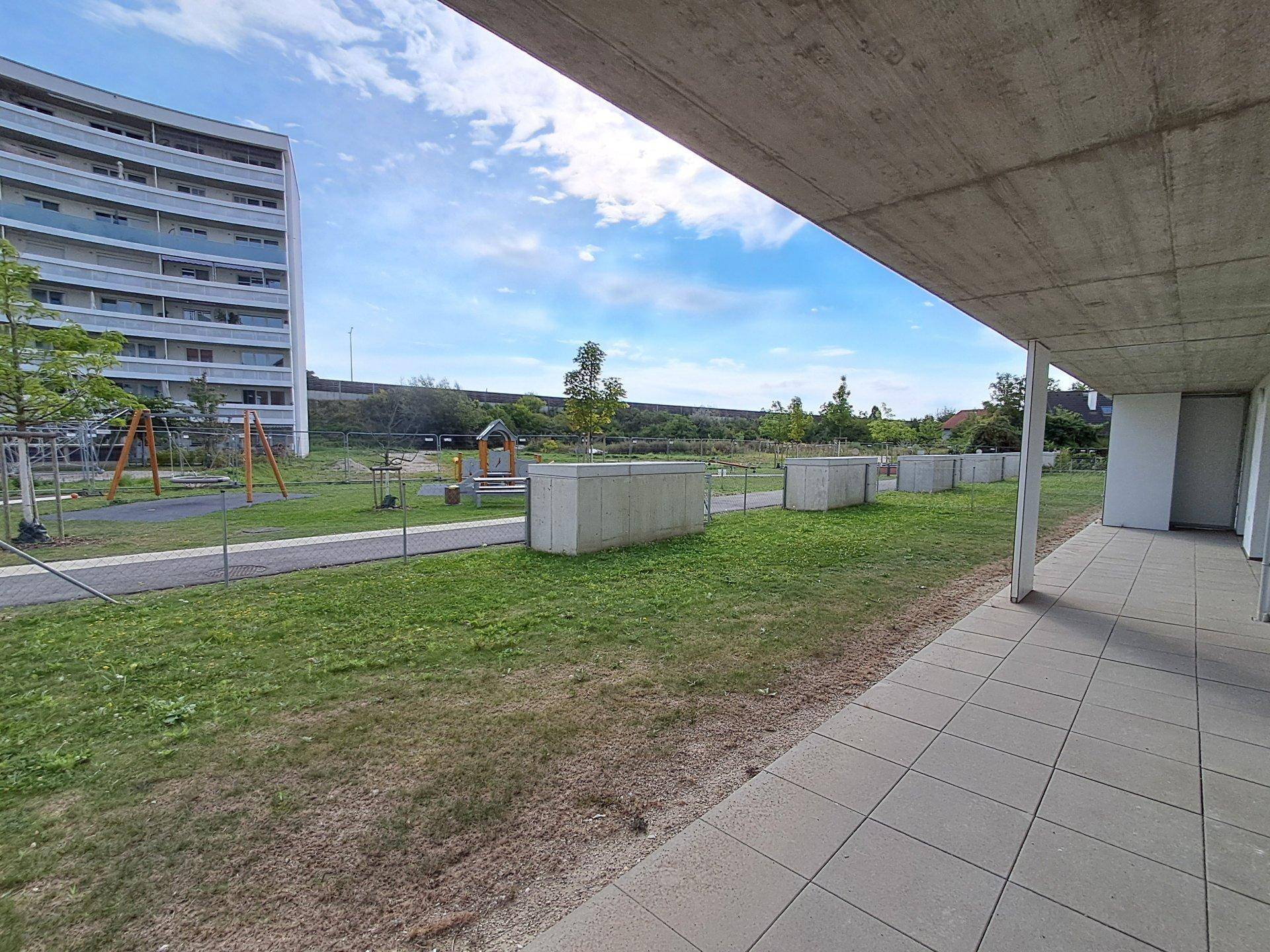 Außenbereich mit gepflasterter Fläche, Rasen und einem Spielplatz, umgeben von moderner Architektur.