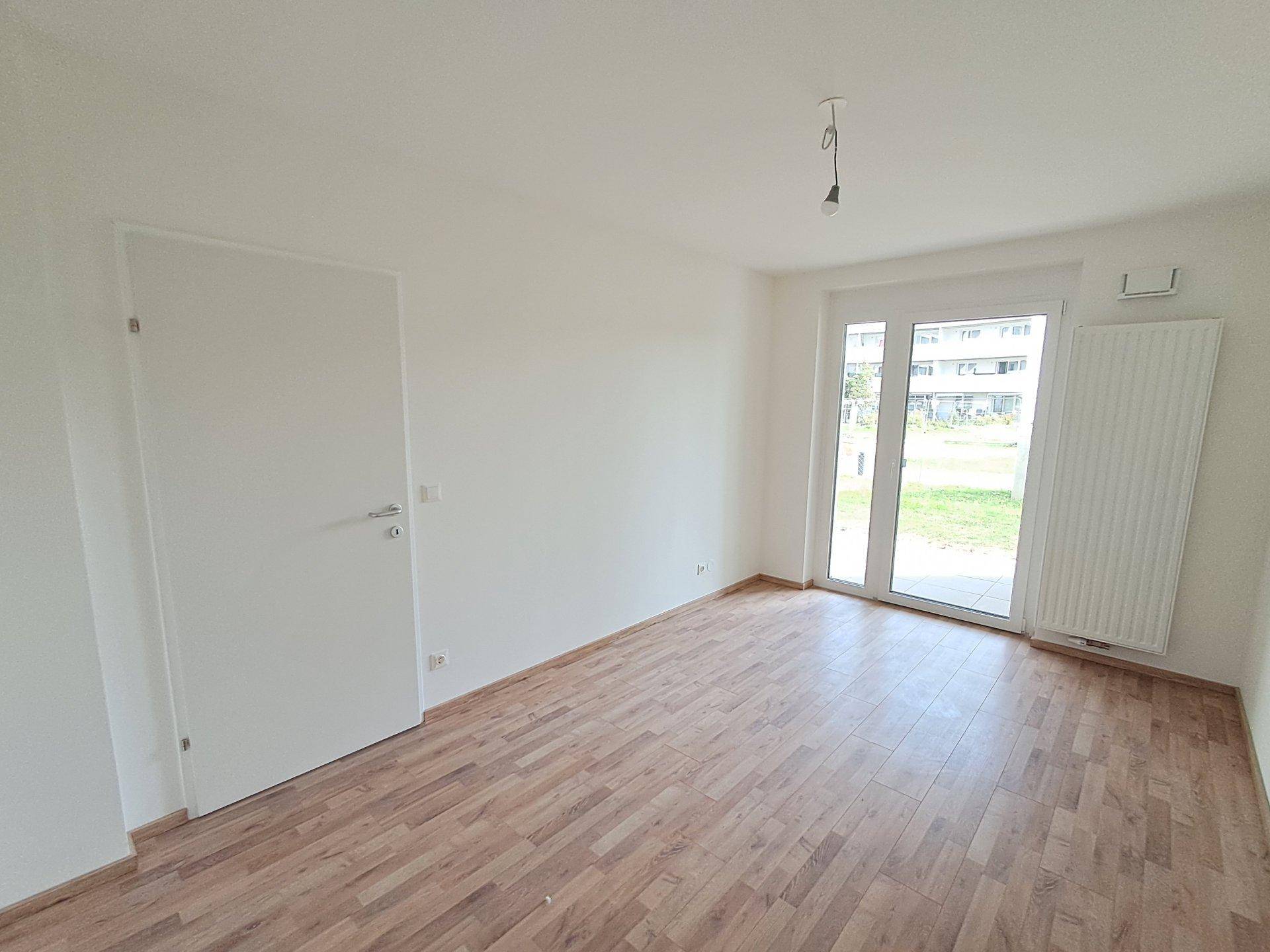 Heller, leerer Raum mit Holzoptikboden und Zugang zu einem sonnigen Außenbereich.