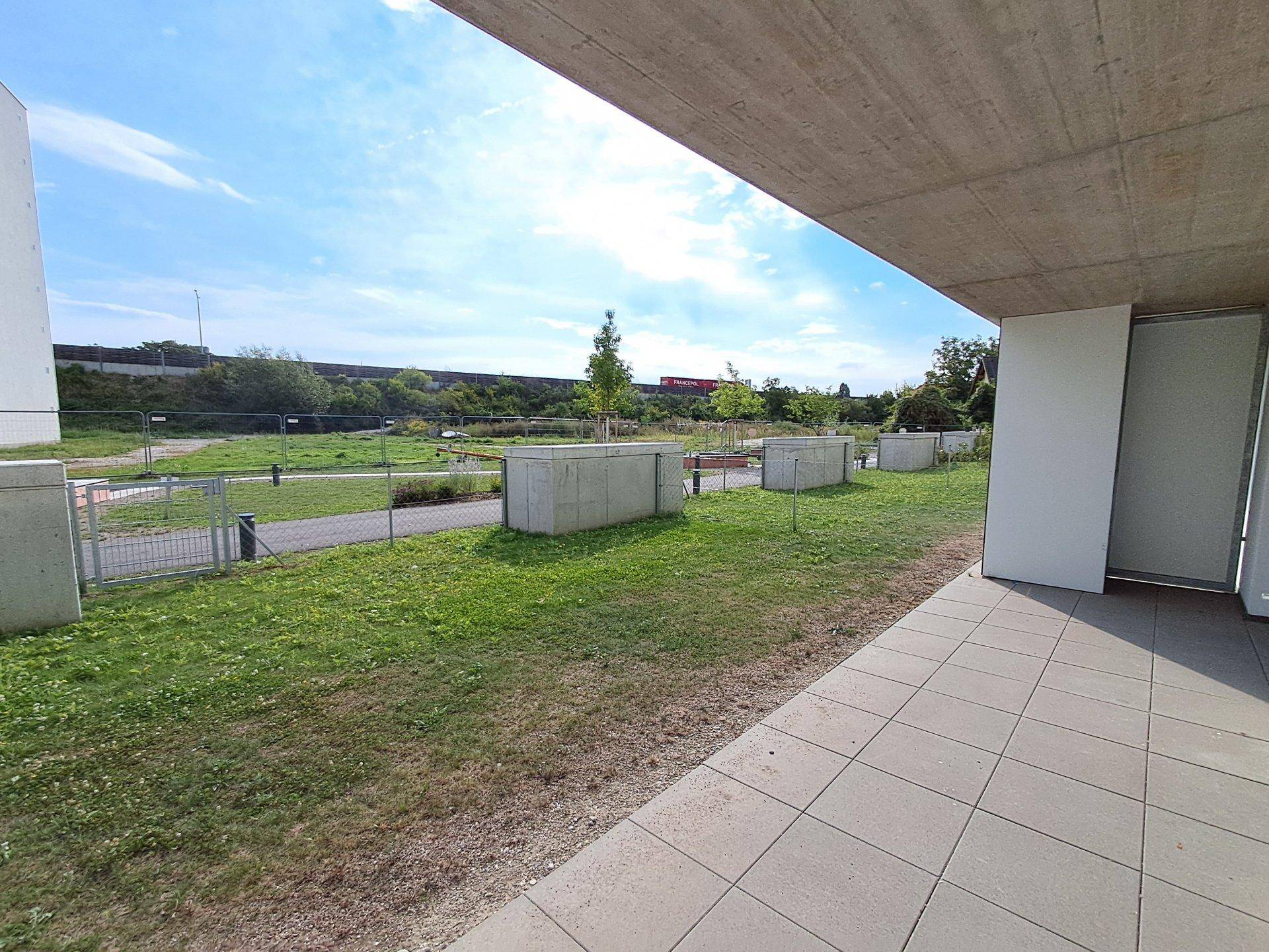 Außenbereich mit gepflasterter Terrasse und gemeinschaftlicher Grünfläche unter blauem Himmel.