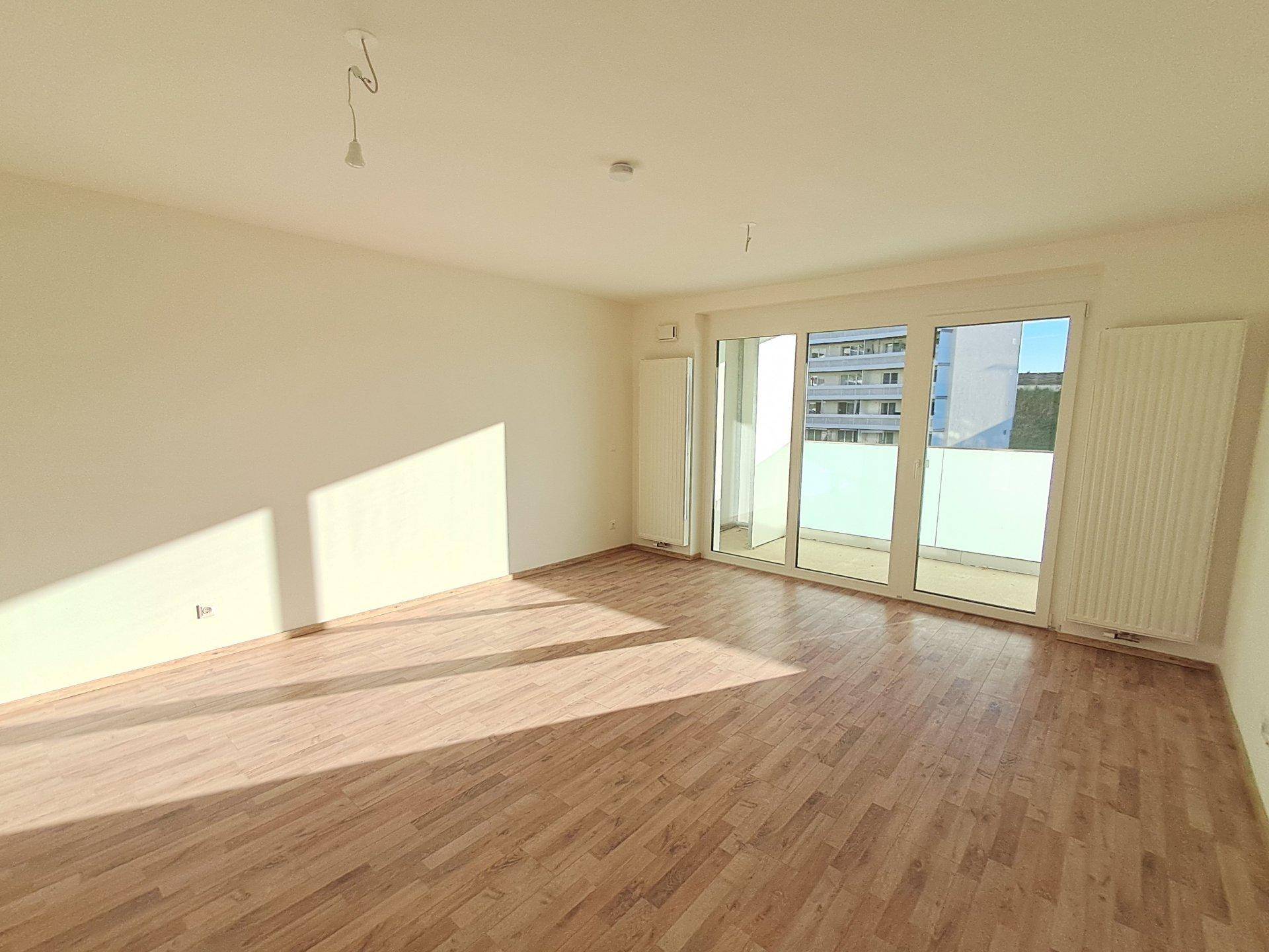 Helles, leeres Wohnzimmer mit Laminatboden und großen Fenstern, die viel Sonnenlicht hereinlassen und Zugang zum Balkon bieten.