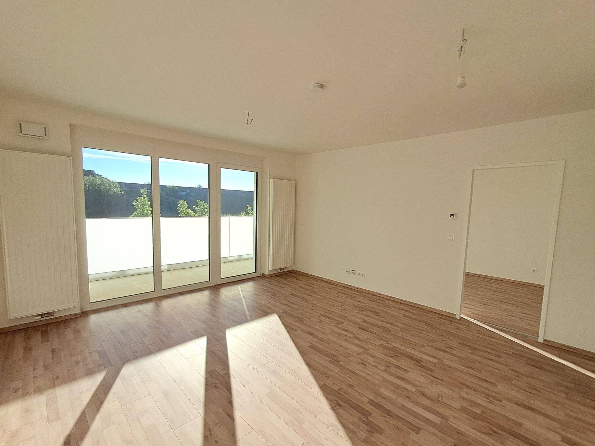 Geräumiges, leeres Wohnzimmer mit Laminatboden und großen Fenstern, die viel Tageslicht und einen Blick ins Grüne bieten.