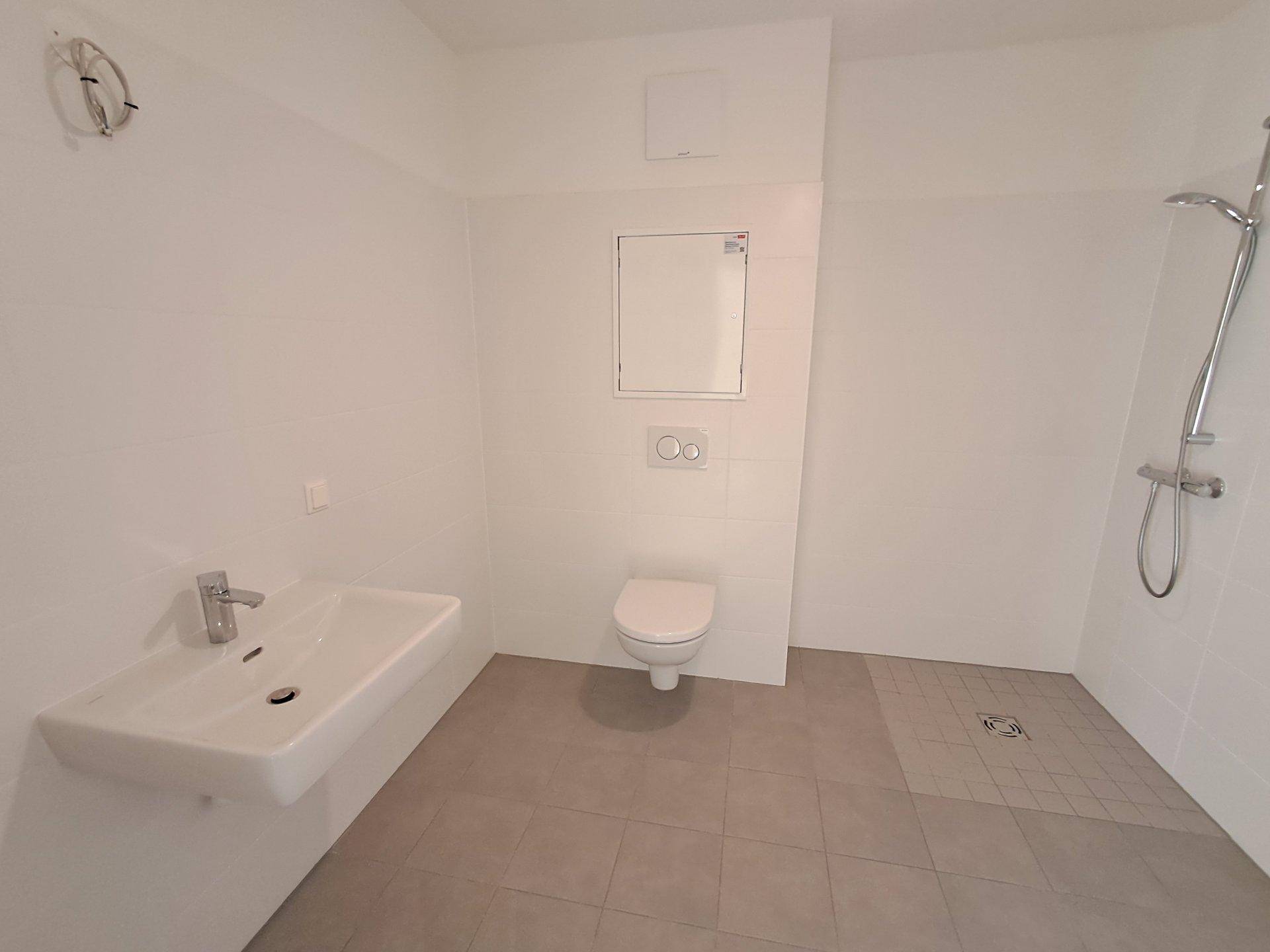 Helles Badezimmer mit weißen Fliesen, Einzelwaschbecken, Wand-WC und ebenerdiger Dusche, unmöbliert.
