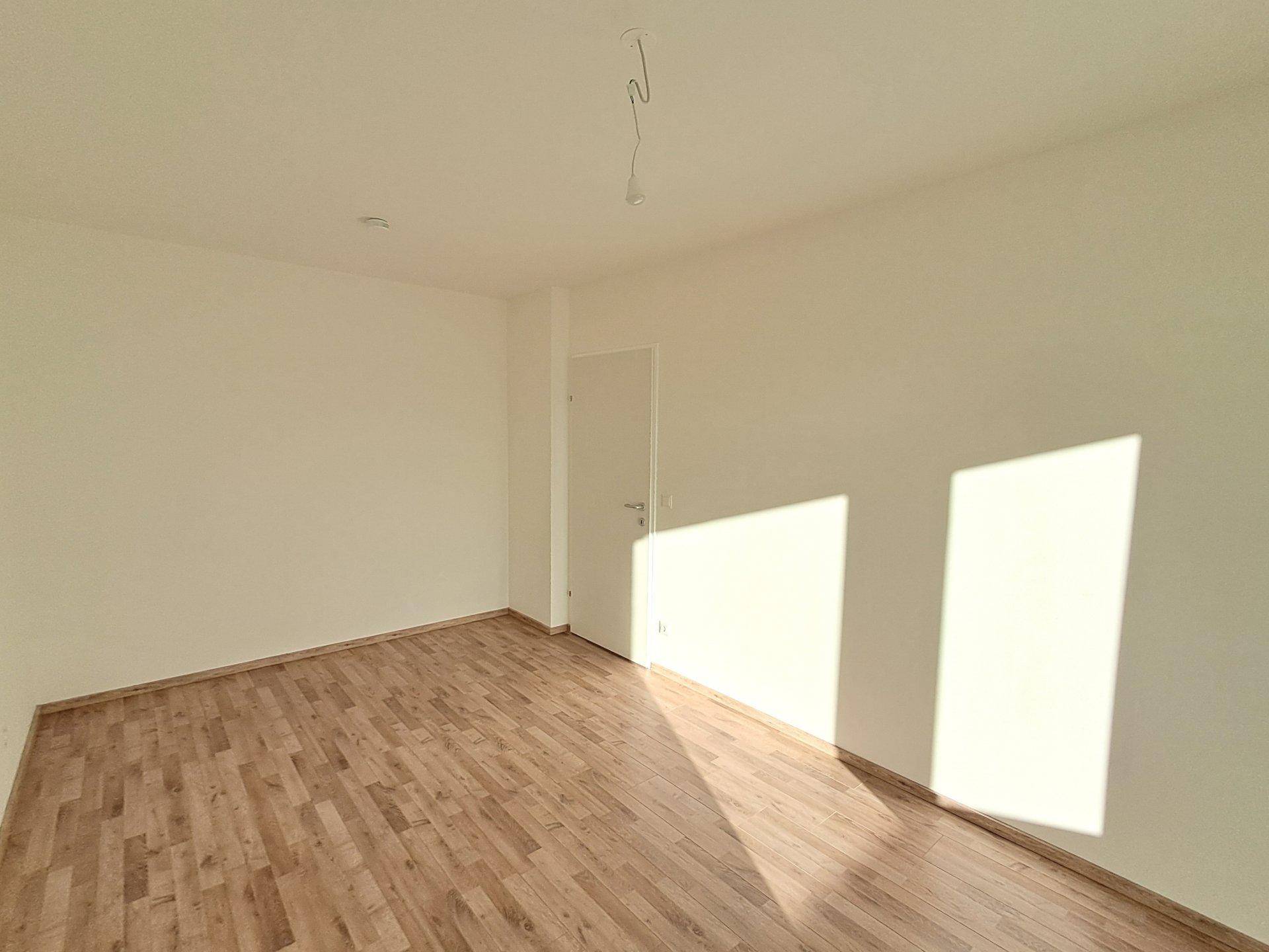 Geräumiges, leeres Zimmer mit Laminatboden und weißen Wänden, das viel Sonnenlicht durch ein nicht sichtbares Fenster erhält.