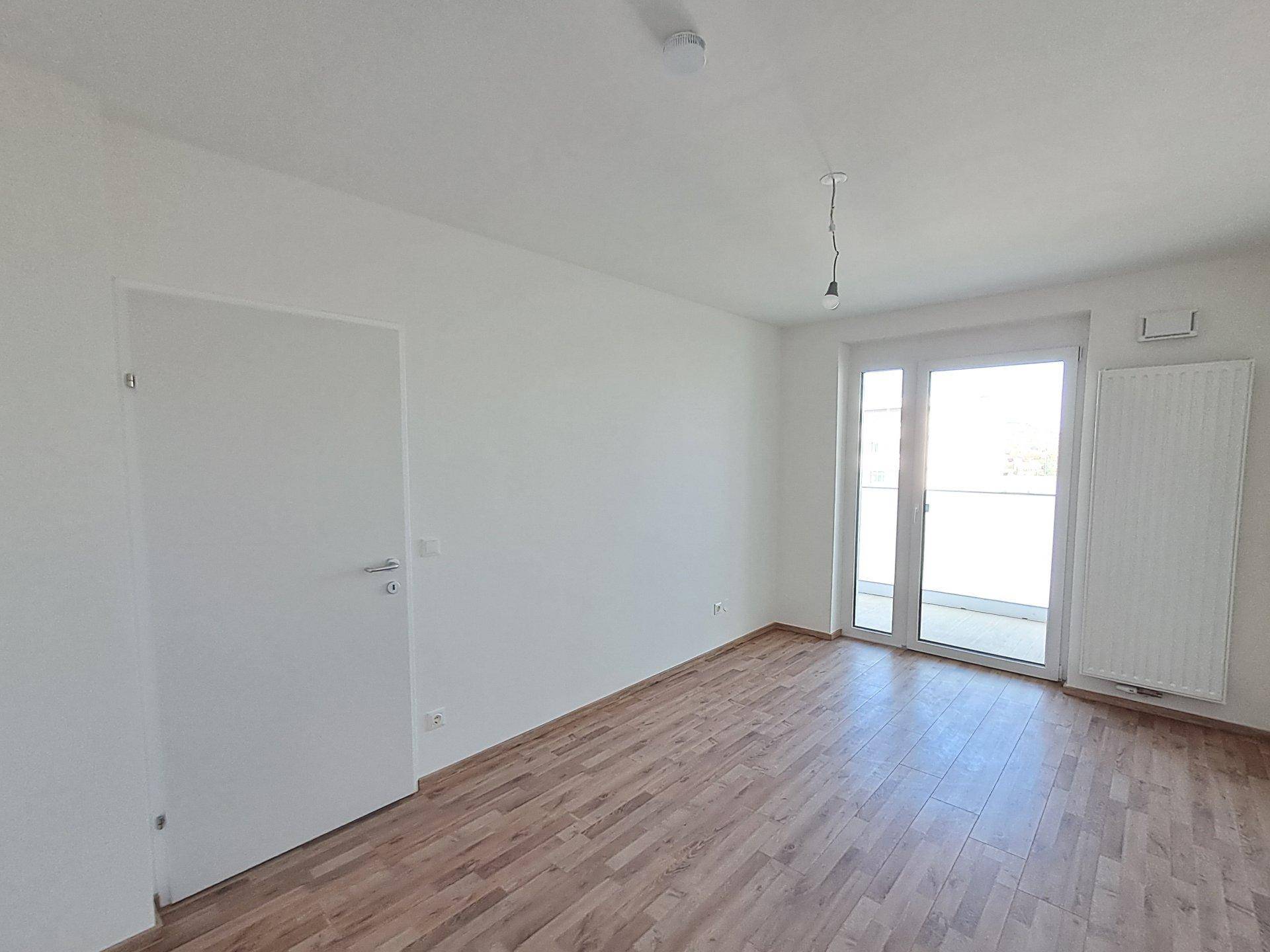 Helles, unmöbliertes Schlafzimmer mit Holzboden und großem Fenster mit Balkonzugang.