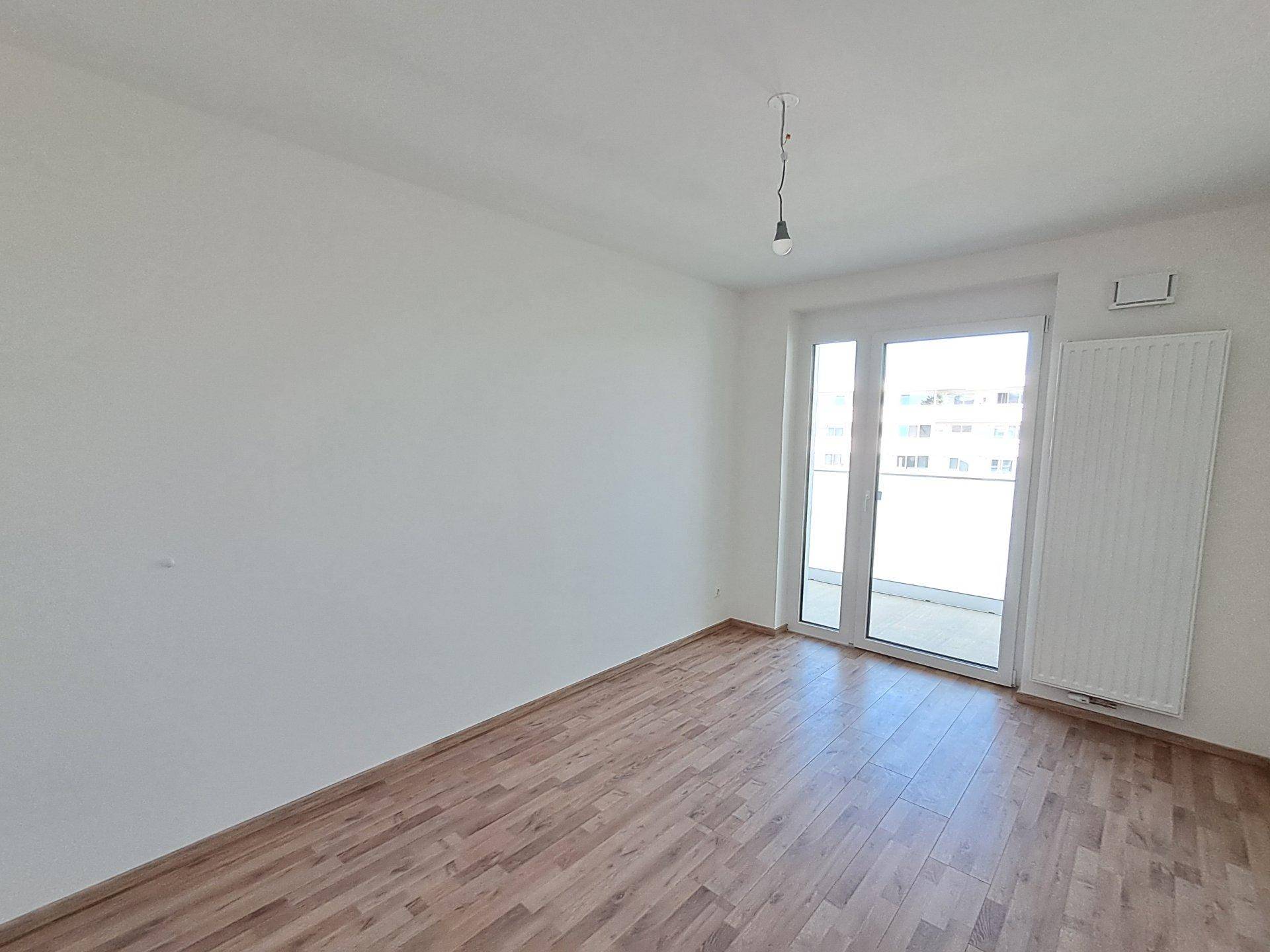 Helles Zimmer mit Laminatboden und breiter Fensterfront, die Zugang zum Balkon bietet.