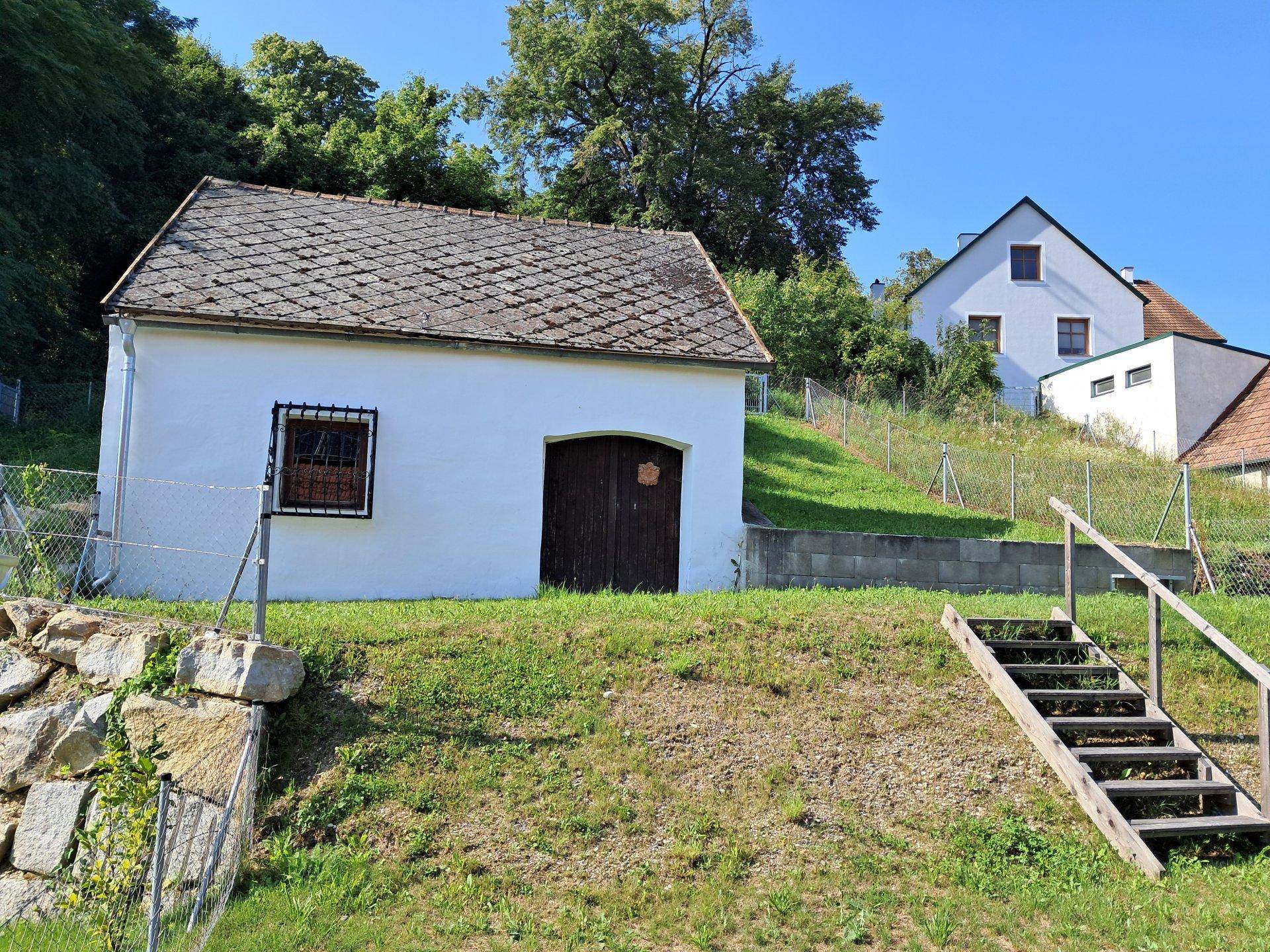 Kleines weißes Nebengebäude mit Holztür und Ziegeldach auf einem grünen Hügel.