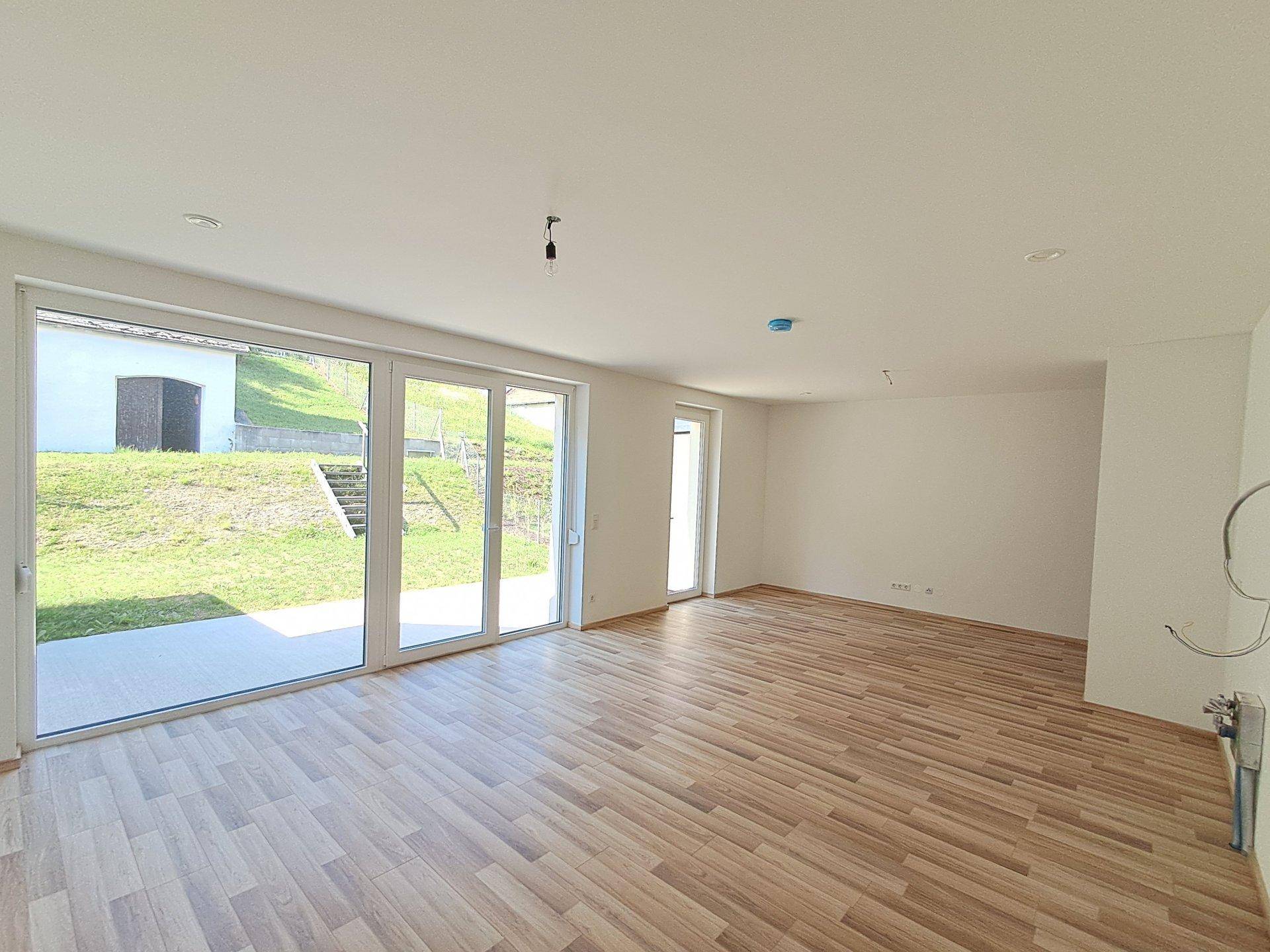 Heller, leerer Wohnraum mit Holzfußboden und großen Glasschiebetüren zur Terrasse.