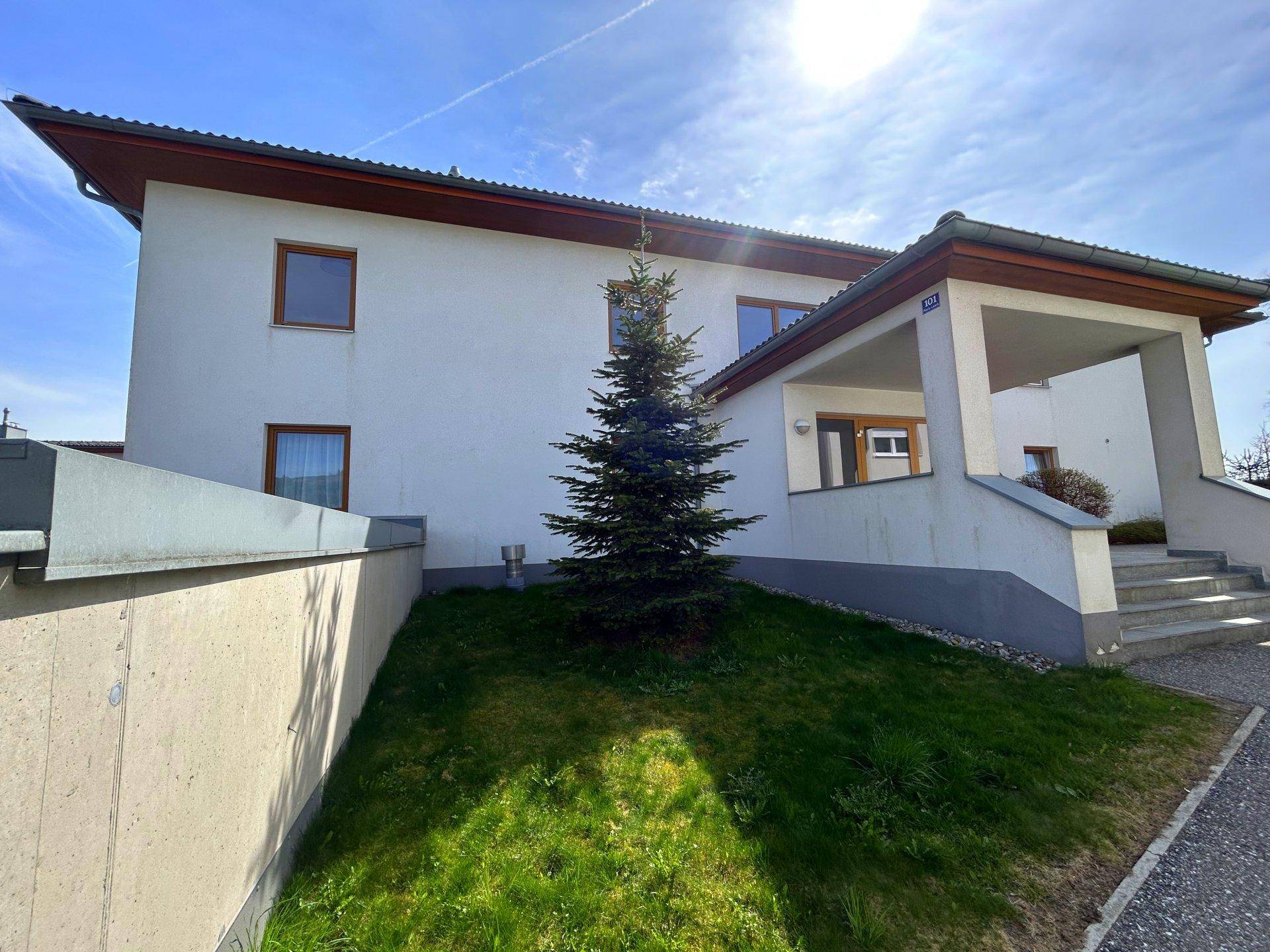 Gebäudeseite mit Außentreppe, überdachtem Eingang und einem Nadelbaum im gepflegten Vorgarten.