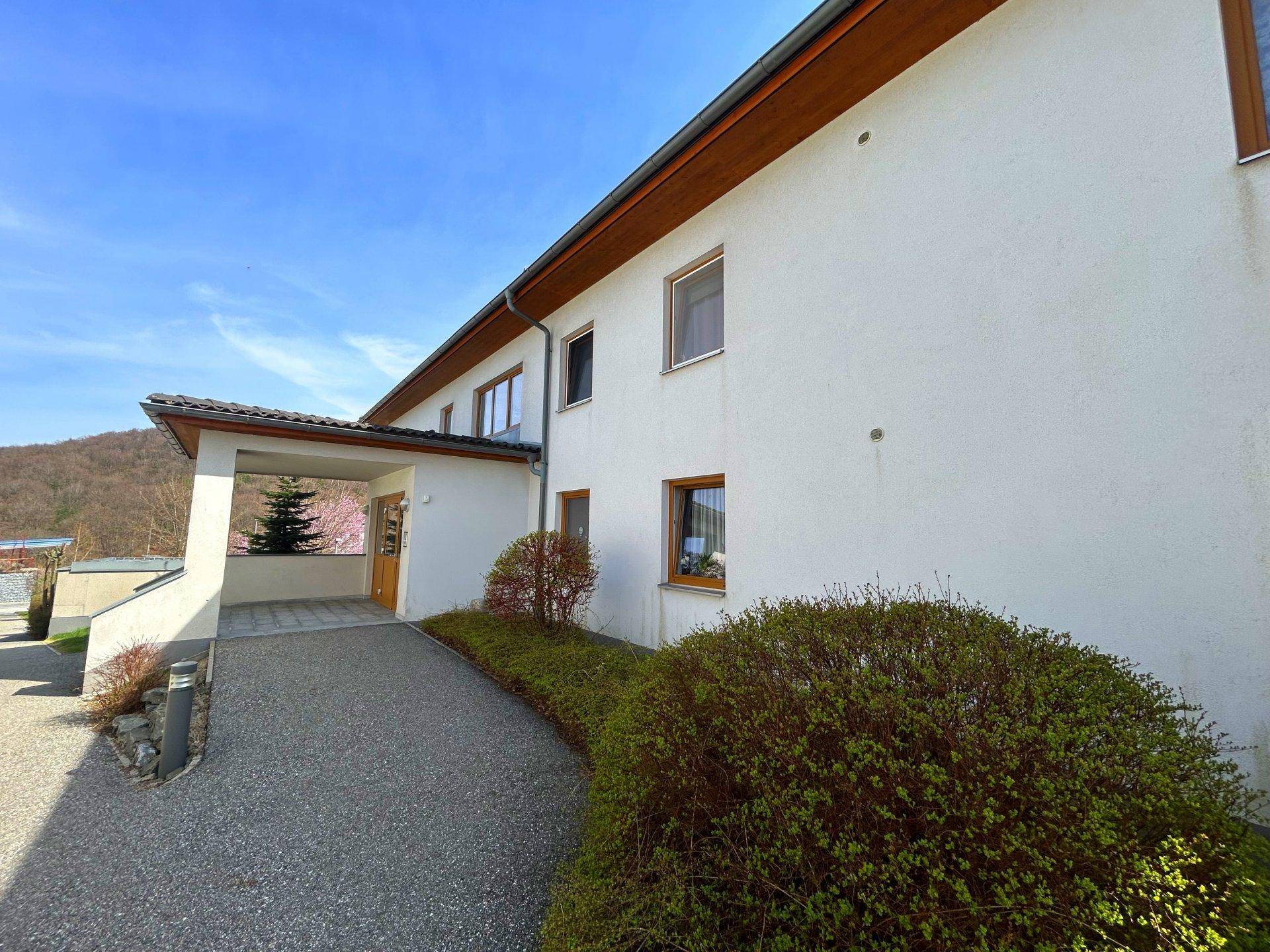 Seitenansicht des Gebäudes mit überdachtem Eingangsbereich und gepflegten Büschen im Vorgarten.