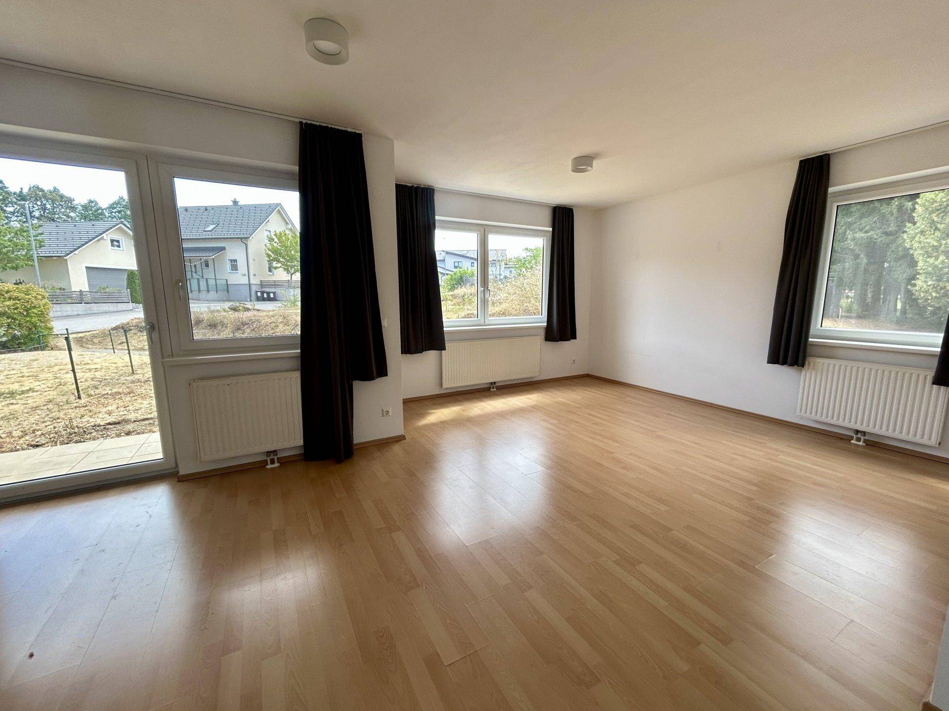 Helles, leeres Wohnzimmer mit hellem Laminatboden und großen Fenstern mit Blick ins Grüne.