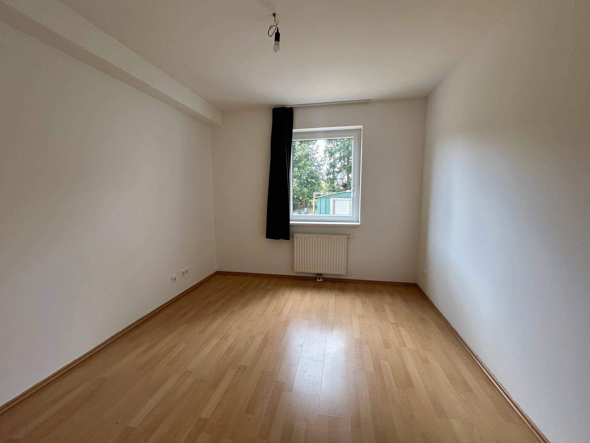 Geräumiges Zimmer mit Laminatboden, weiß gestrichenen Wänden und einem Fenster mit Blick ins Grüne.