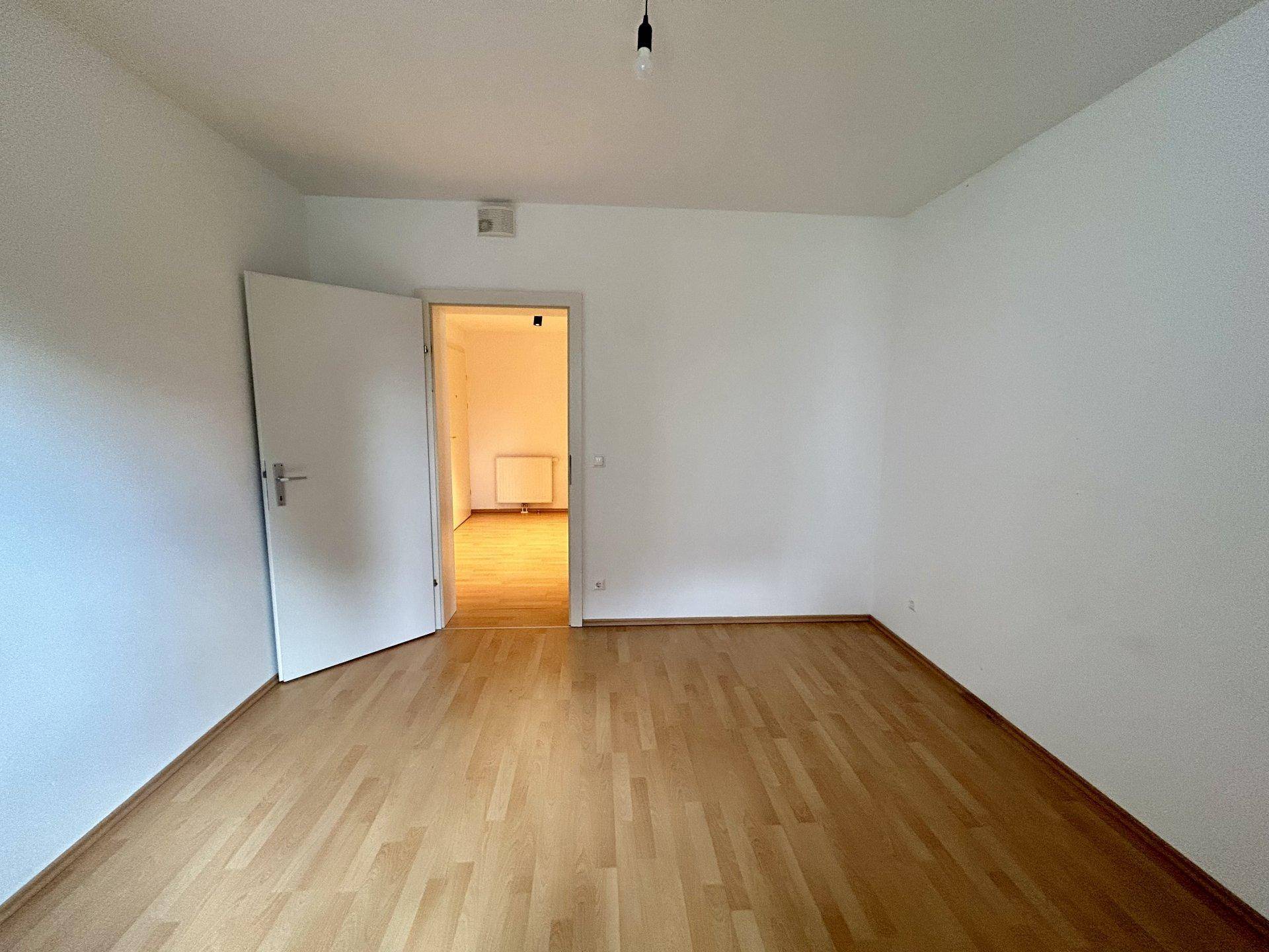 Leeres Zimmer mit hellem Laminatboden und weiß gestrichenen Wänden, ideal als Schlaf- oder Arbeitszimmer.
