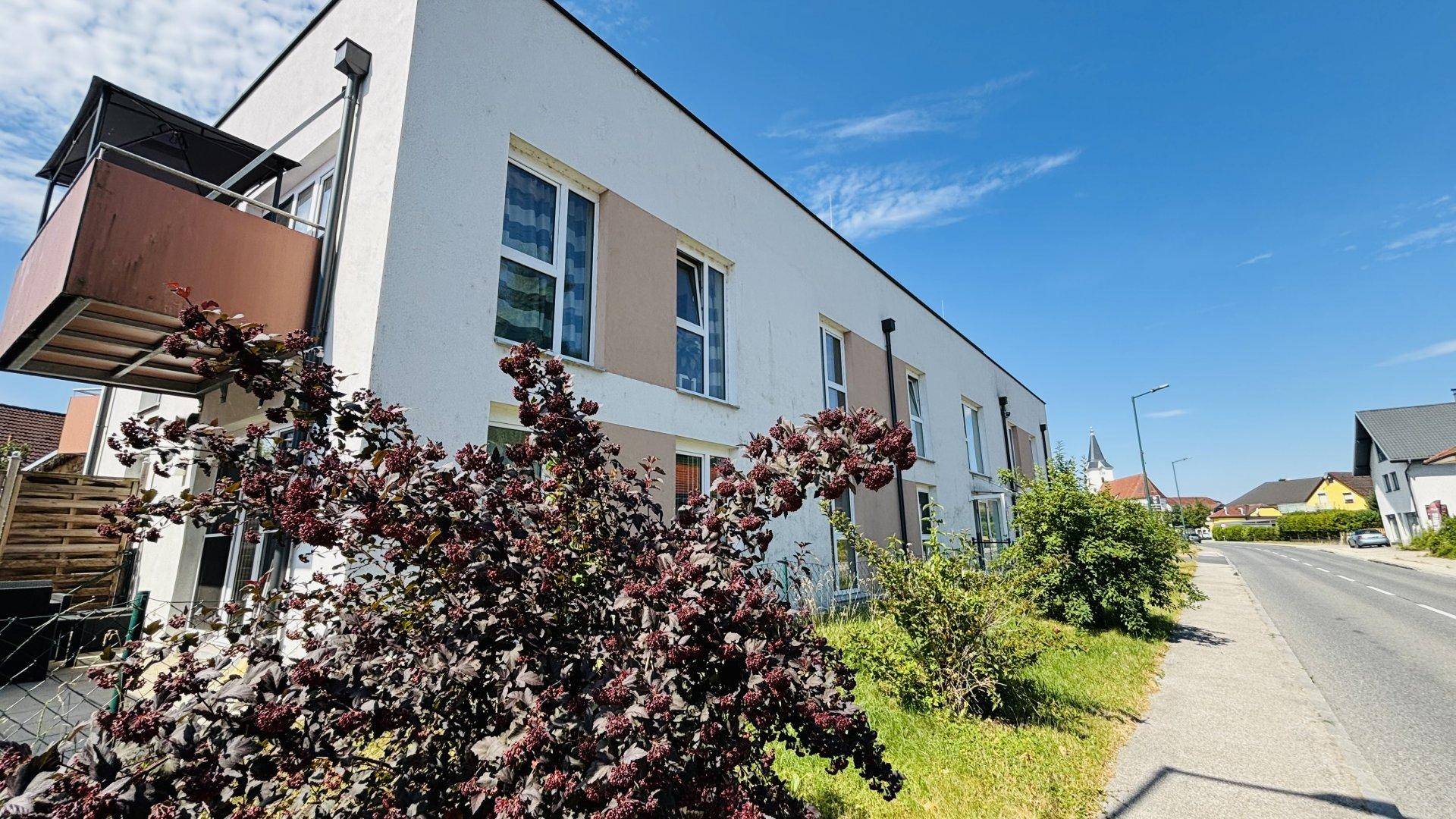 Seitenansicht eines modernen Gebäudes mit Balkon, umgeben von Grünflächen und einer ruhigen Straße.
