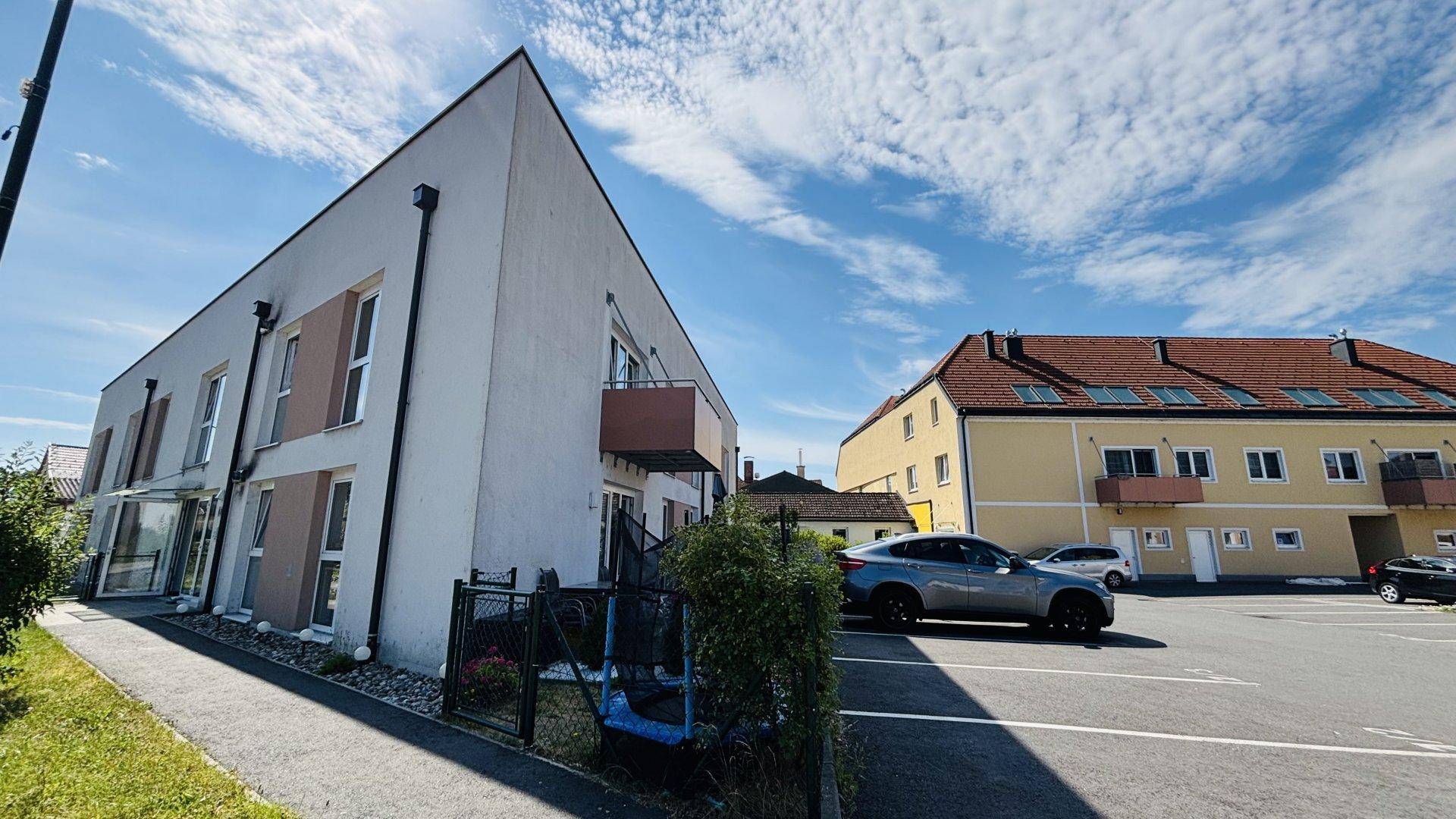 Modernes Wohngebäude mit heller Fassade und Parkplätzen, umgeben von weiteren Gebäuden und Grünflächen.