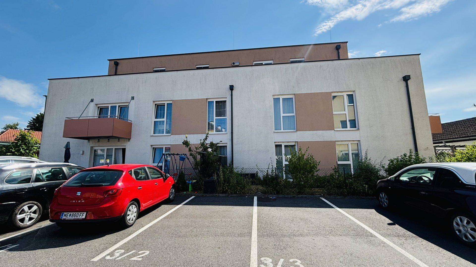 Vorderansicht eines modernen Wohnhauses mit heller Fassade, Fenstern und Parkplätzen im Sonnenschein.
