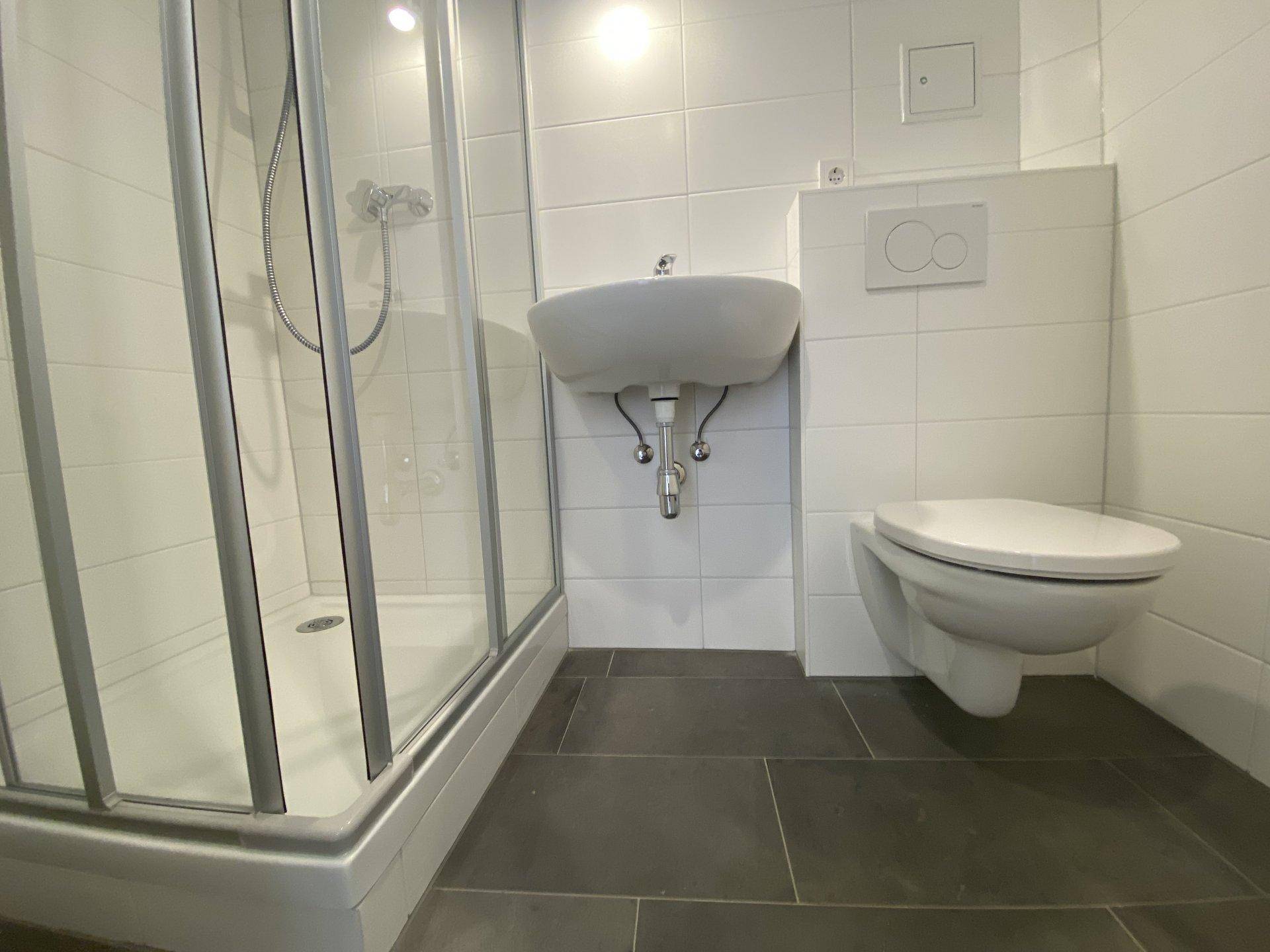 Modernes Badezimmer mit weiß gefliesten Wänden, dunklem Boden, Duschkabine und wandhängendem WC.