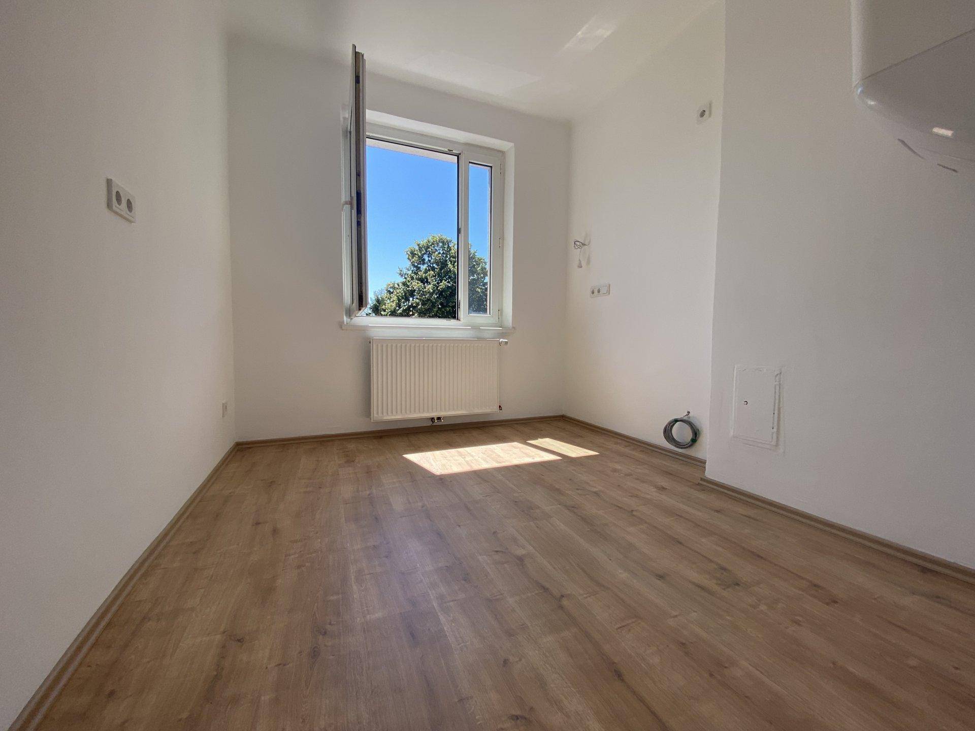 Leerer Raum mit Holzboden und großem Fenster, das viel Tageslicht hereinlässt und einen Blick ins Grüne bietet.