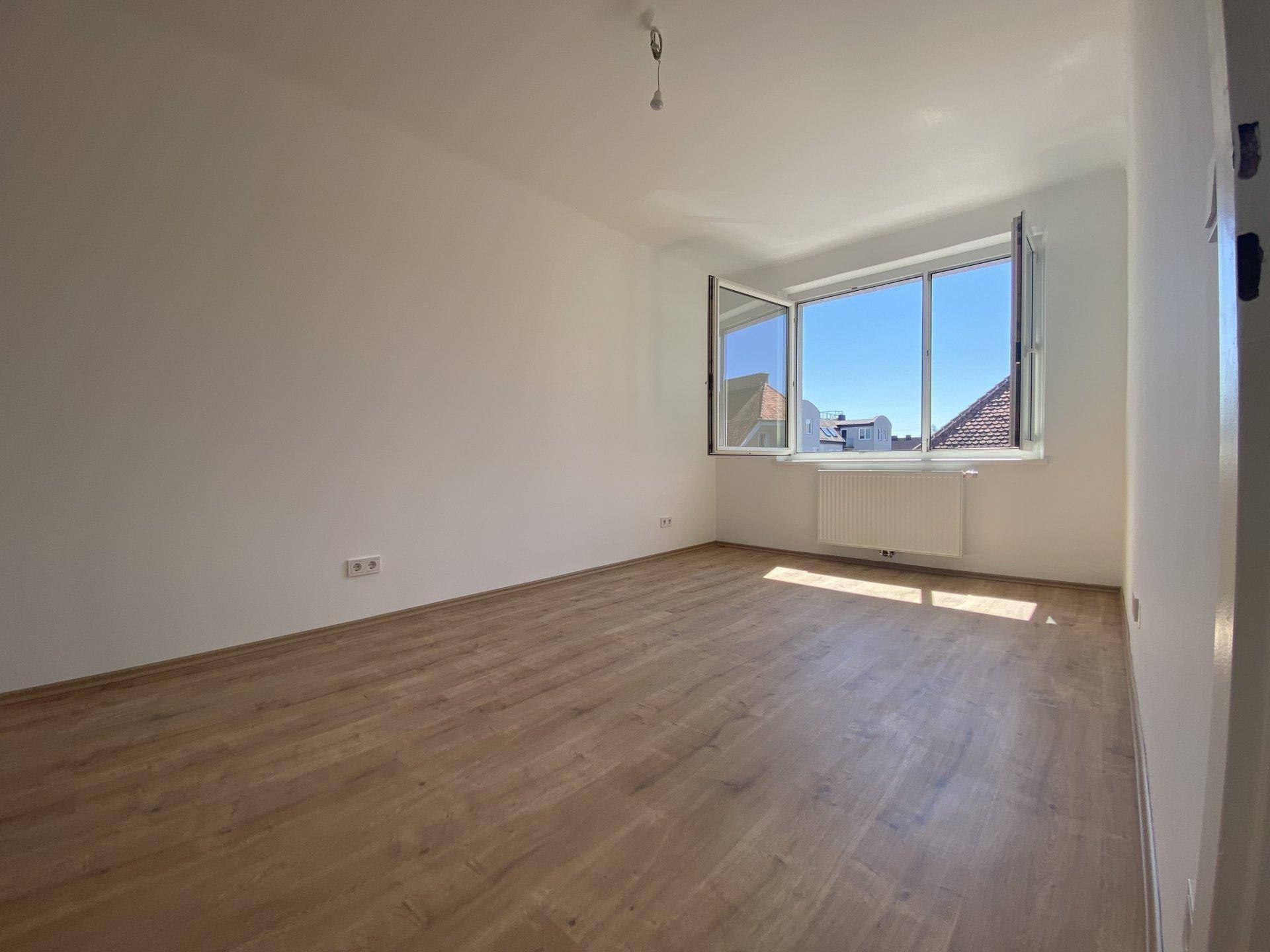 Heller, leerer Raum mit Holzboden und großem Fenster, das einen weiten Blick über die Dächer ermöglicht.