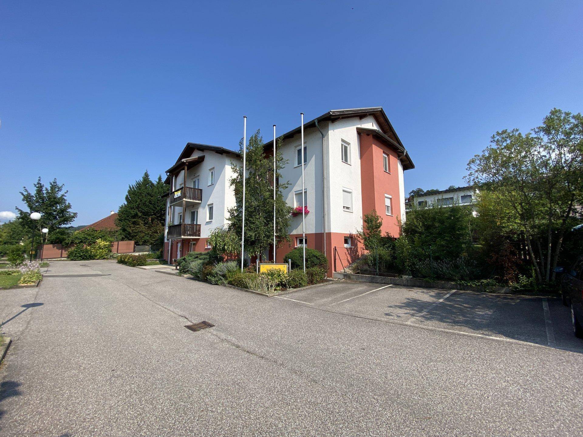 Mehrfamilienhaus mit Parkplätzen und gepflegter Grünanlage unter strahlend blauem Himmel.