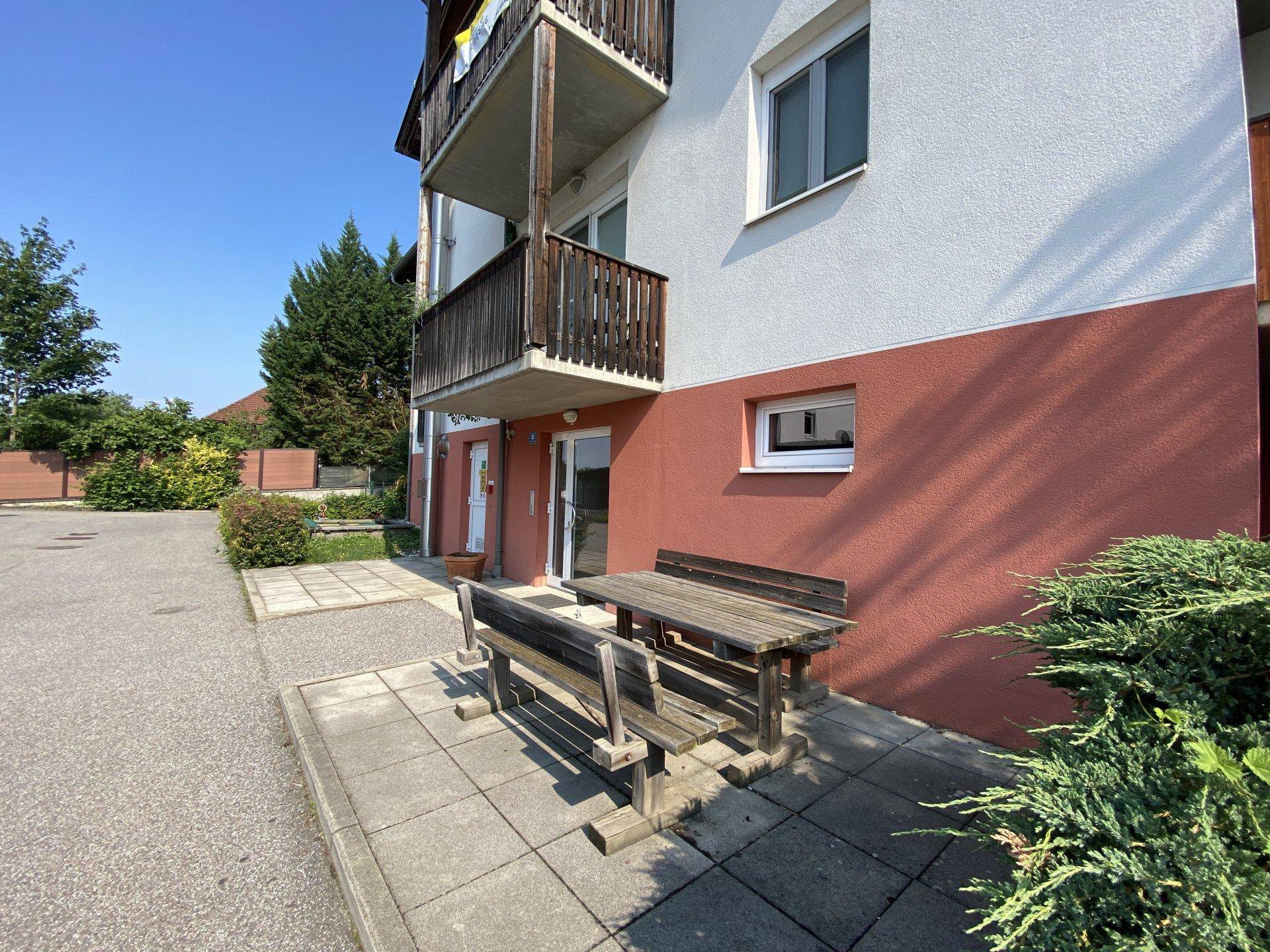 Eingangsbereich eines Gebäudes mit Holztisch und Bänken sowie einem Balkon im Obergeschoss.