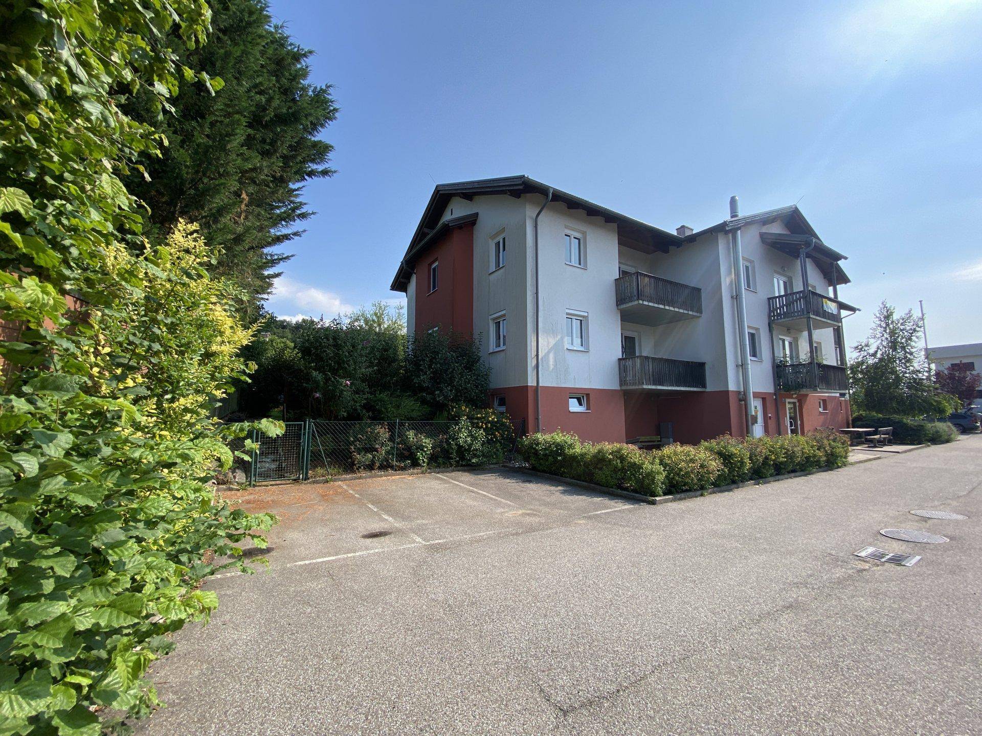 Seitenansicht eines Mehrfamilienhauses mit mehreren Balkonen und einem Parkplatz.