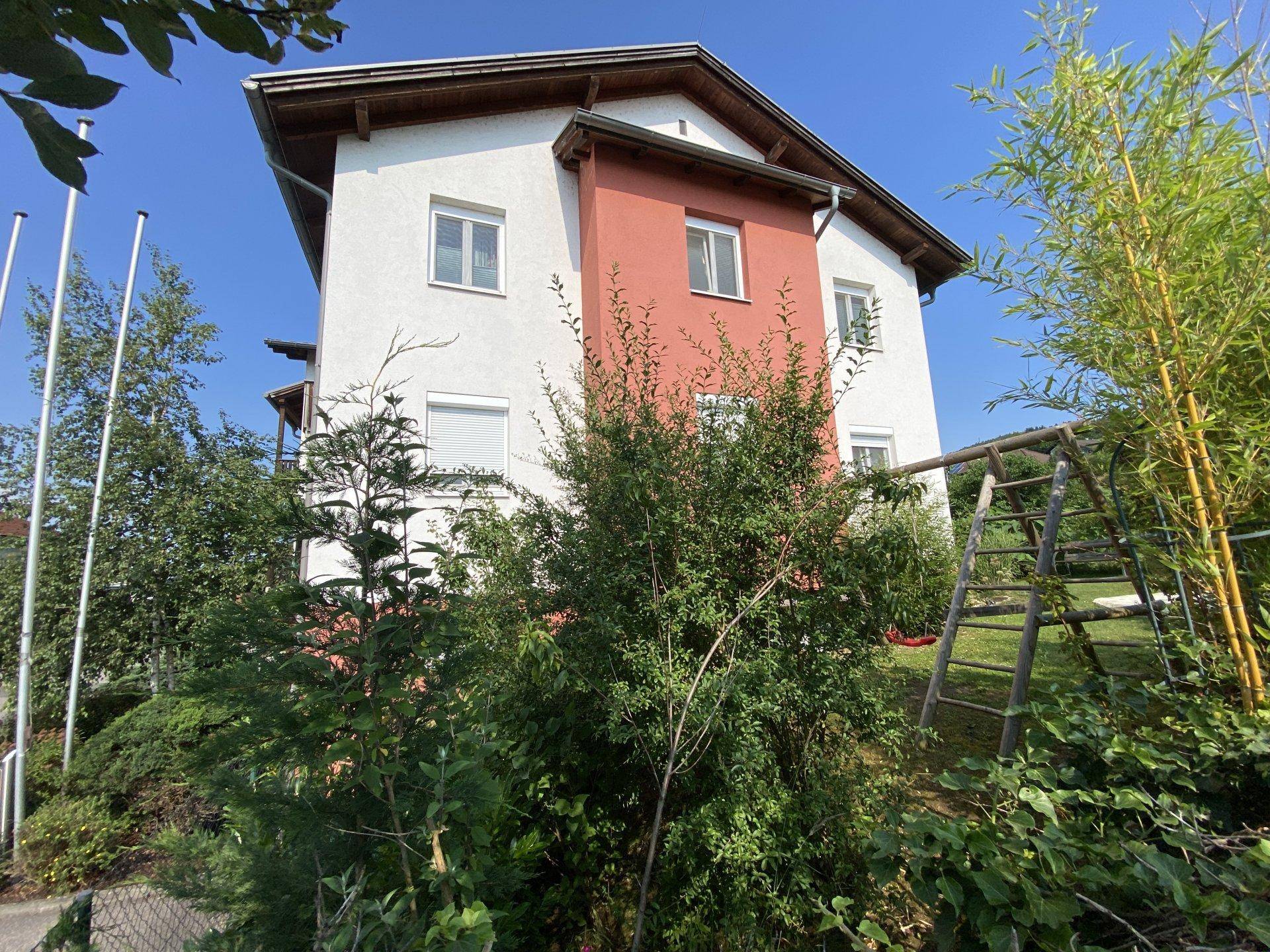 Seitenansicht eines Wohnhauses mit weißer und roter Fassade, umgeben von Sträuchern.
