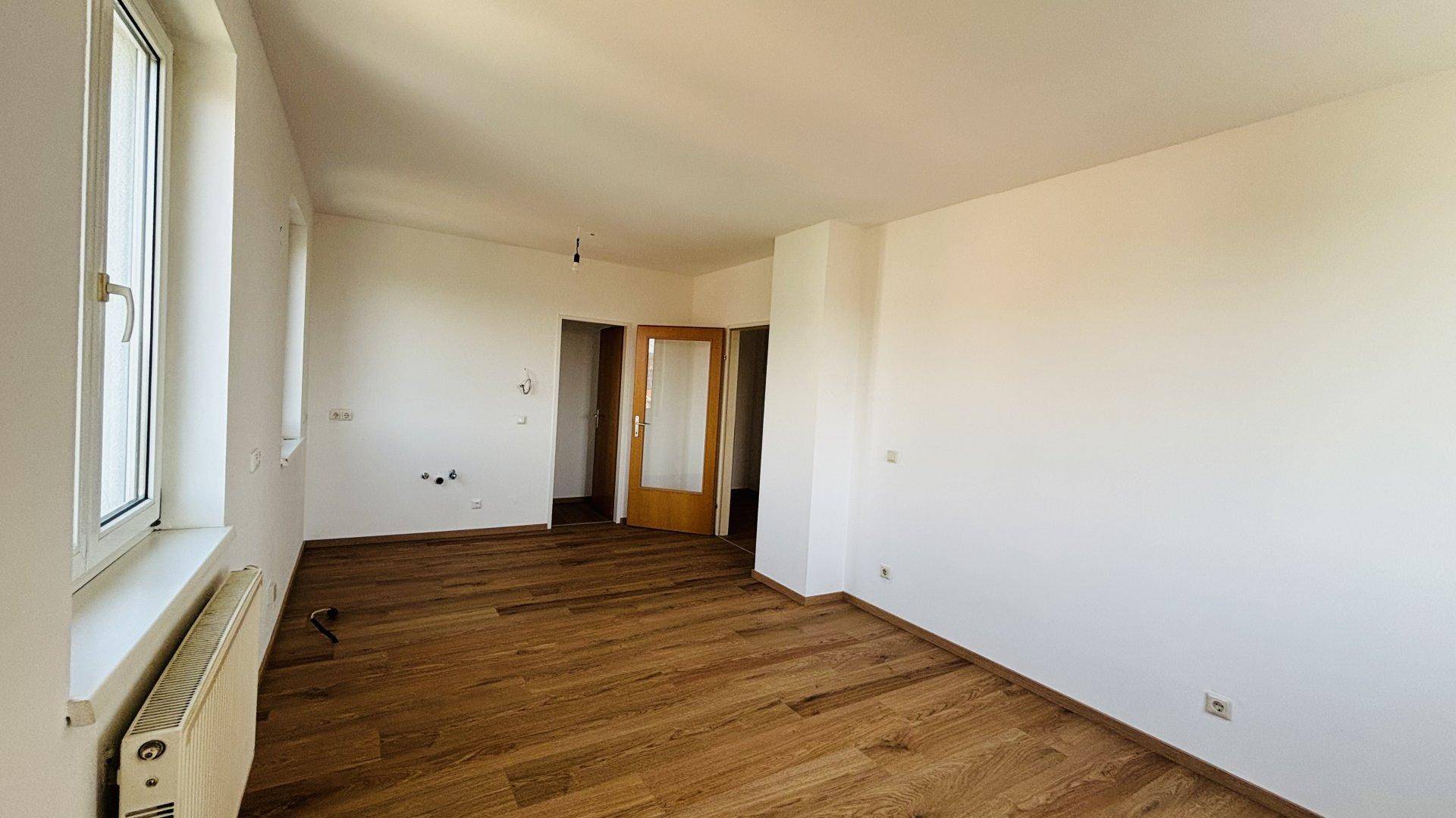 Heller Wohnraum mit Holzfußboden, weißen Wänden und einer Holztür, die zu einem anderen Zimmer führt.