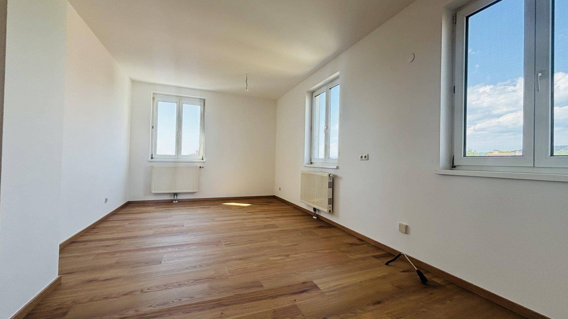 Helles, leeres Zimmer mit Holzfußboden und mehreren Fenstern, die viel Tageslicht hereinlassen.