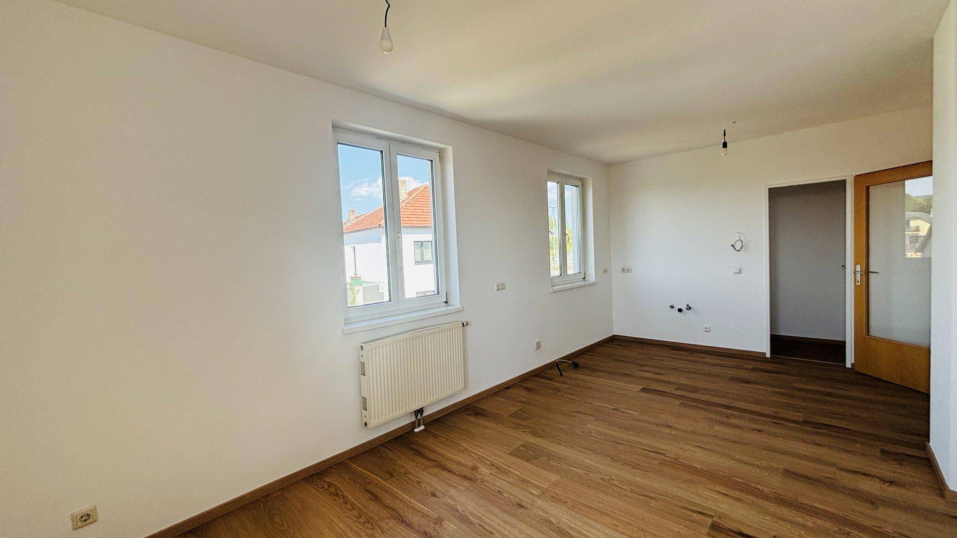 Geräumiges, helles Zimmer mit Holzfußboden, Heizkörpern und einer Tür zu einem weiteren Bereich.