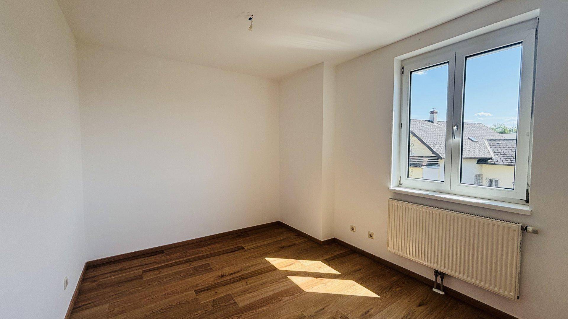 Heller Raum mit Holzfußboden und einem Fenster, das viel Sonnenlicht hereinlässt und eine Aussicht bietet.