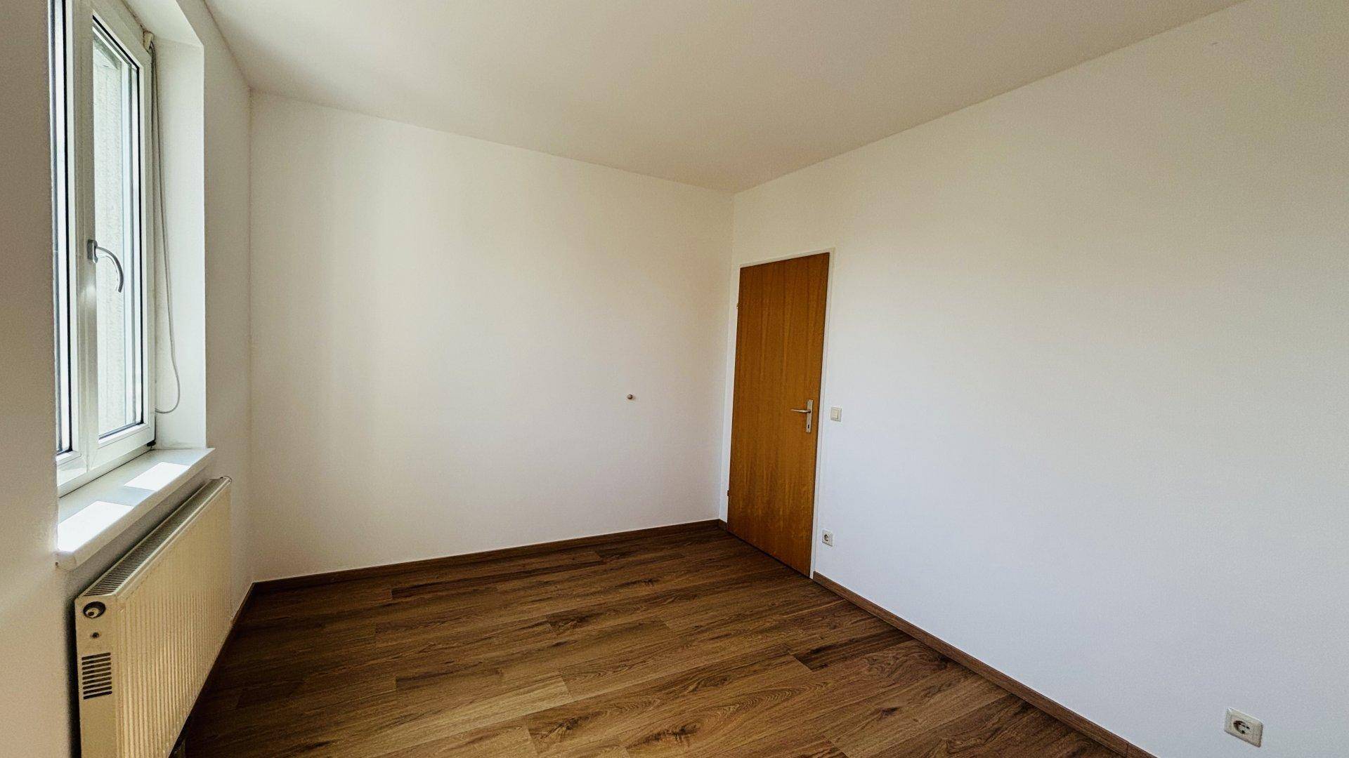 Leeres Zimmer mit Holzfußboden, weiß gestrichenen Wänden und einem Fenster mit Heizkörper.