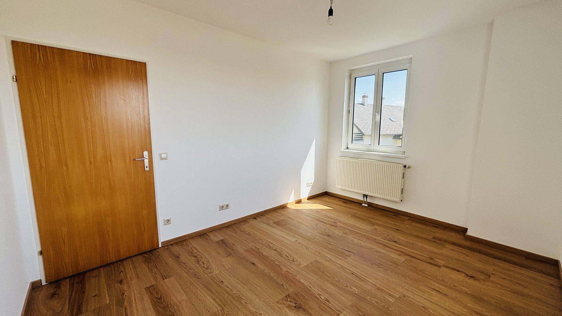 Freundliches Zimmer mit Holzfußboden, weiß gestrichenen Wänden und einer Holztür.