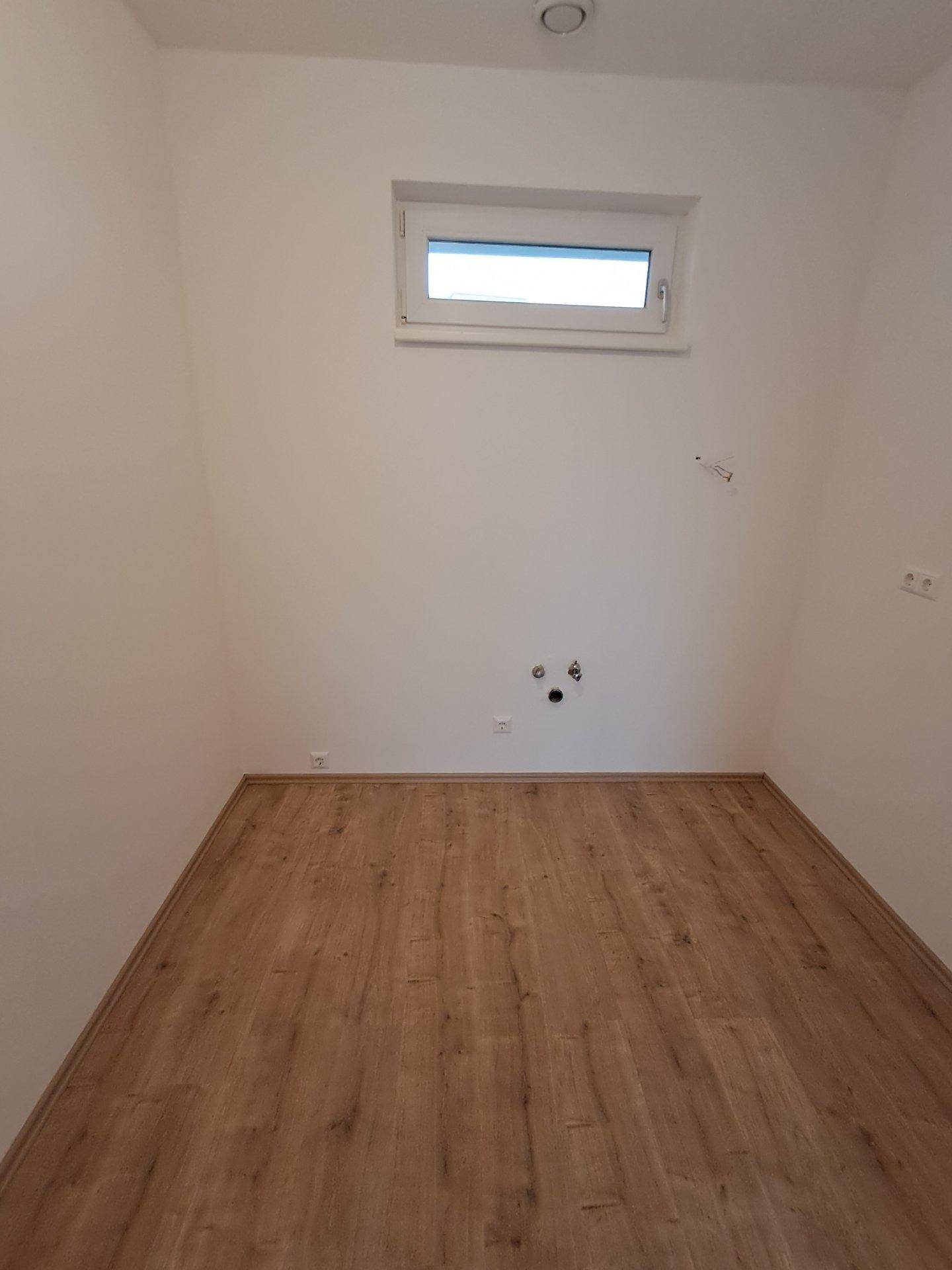 Kleiner, heller Raum mit Fenster, Laminatboden und Anschlüssen für Waschmaschine oder Spüle.