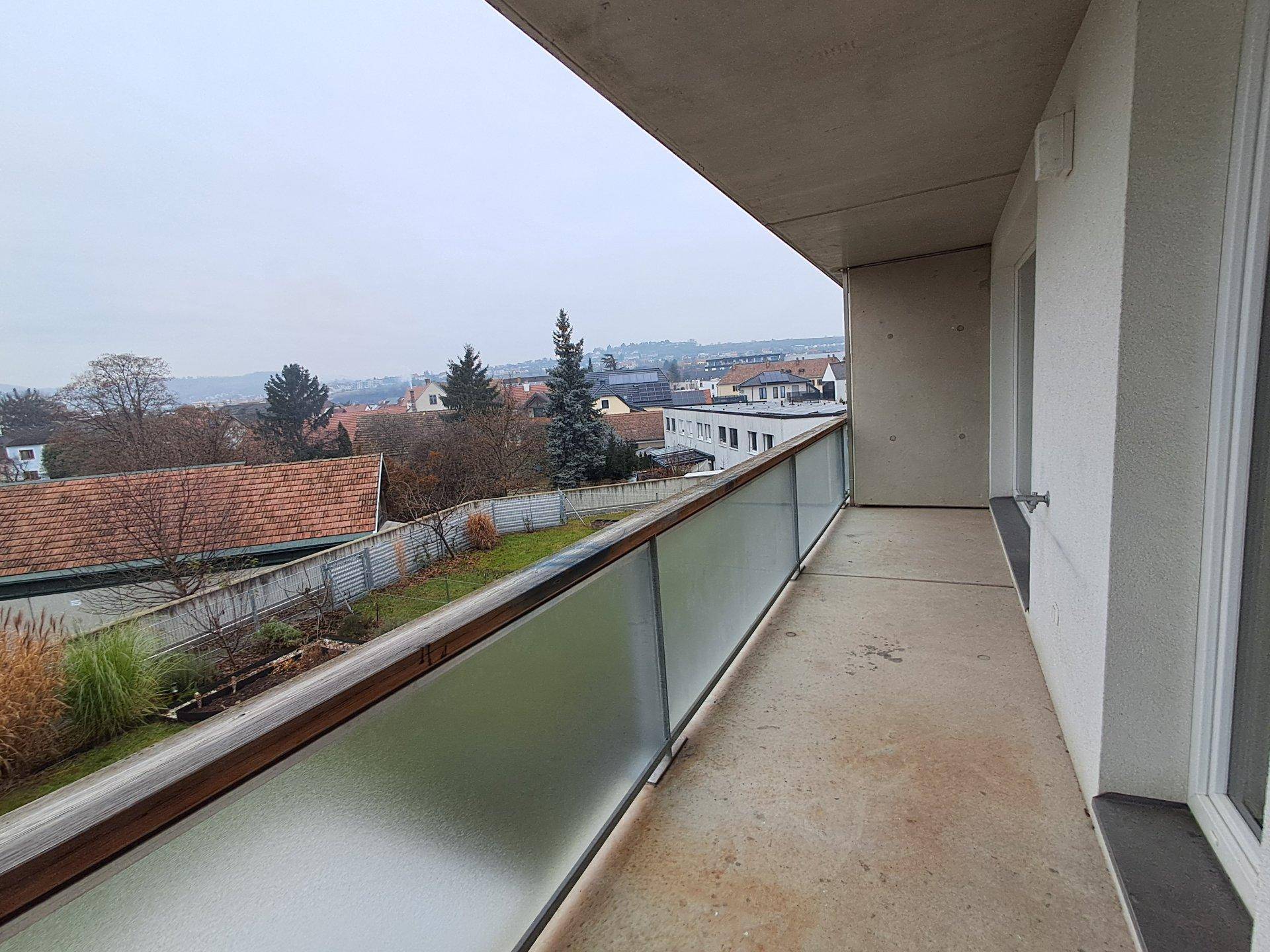 Langer Balkon mit Milchglasbrüstung und Blick über eine Wohnsiedlung mit Bäumen.
