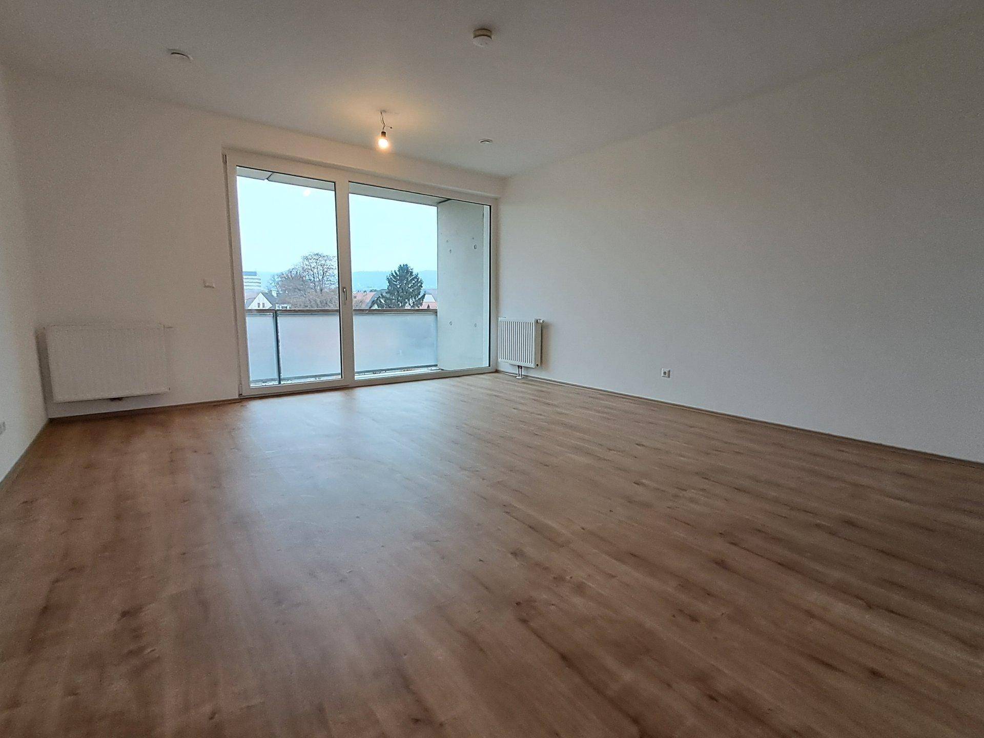 Geräumiger Wohnbereich mit Laminatboden, großen Fenstern und Zugang zu einem Balkon.