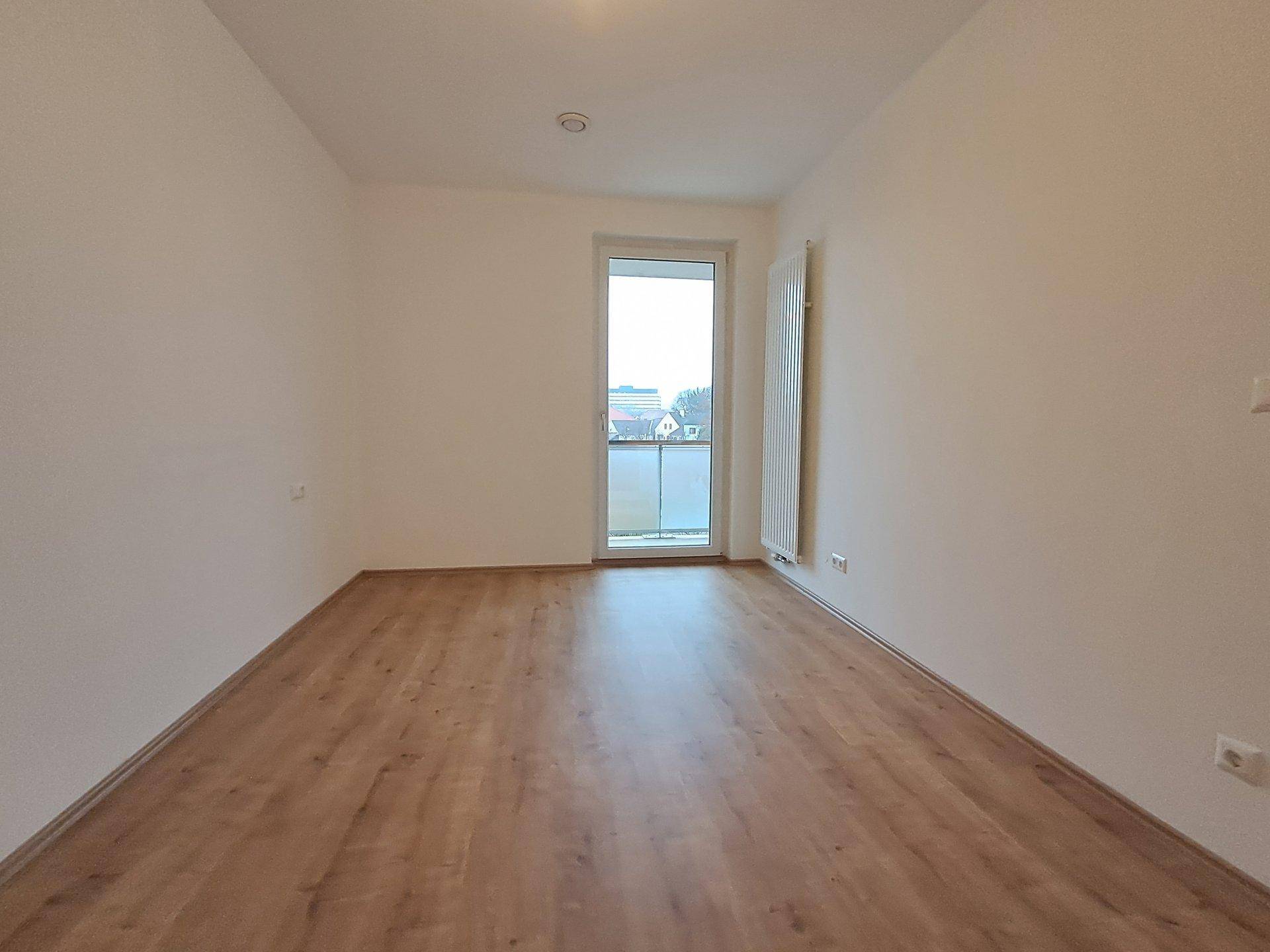 Helles Schlafzimmer mit Laminatboden, weißen Wänden und Zugang zu einem französischen Balkon.