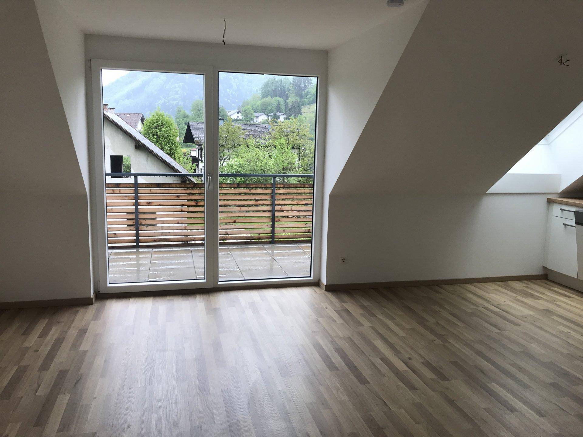Geräumiger Wohnbereich mit Holzfußboden und großen Fenstern, die Zugang zu einem Balkon bieten.