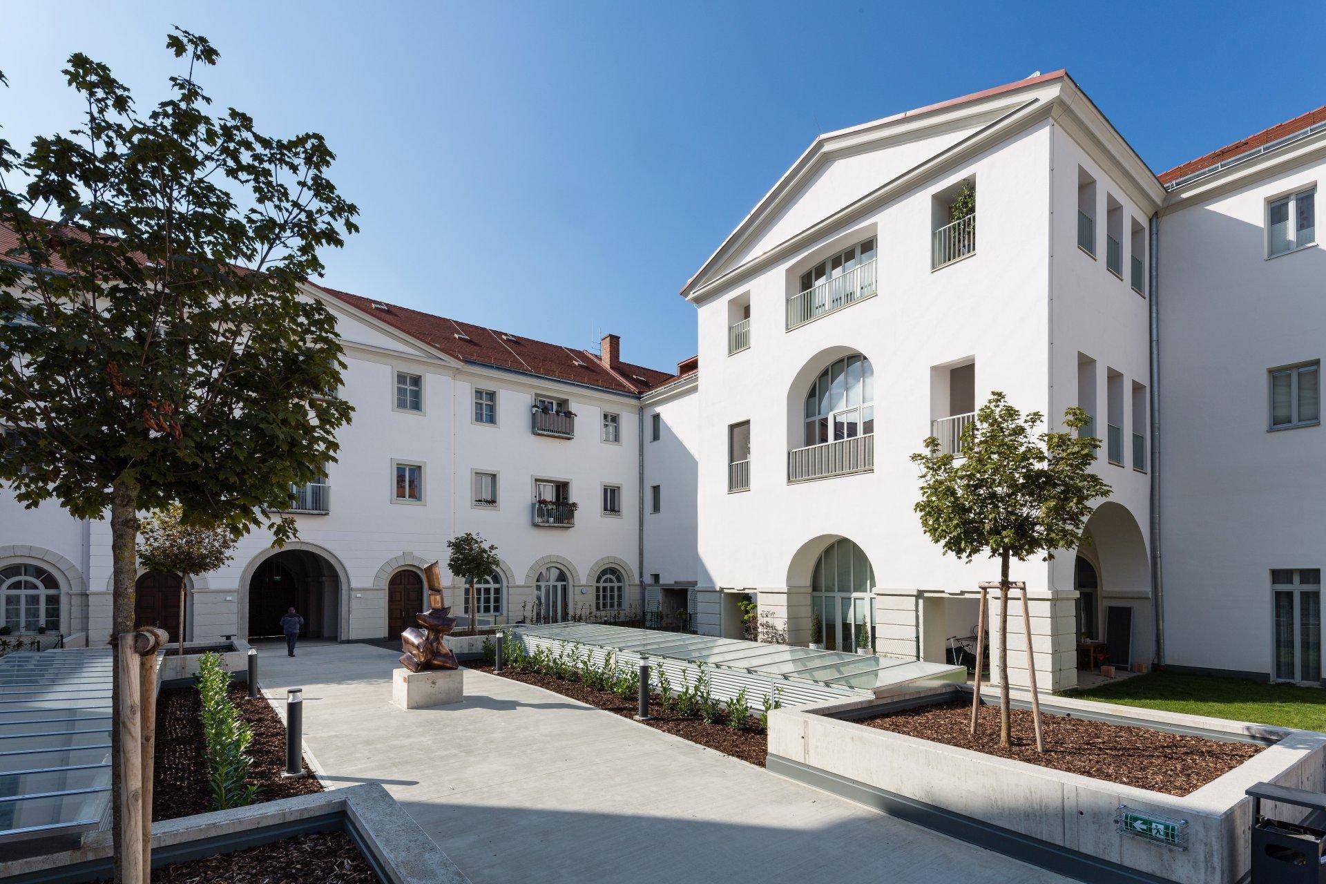 Heller Innenhof eines modernen Gebäudekomplexes mit Bäumen und gepflasterten Wegen.