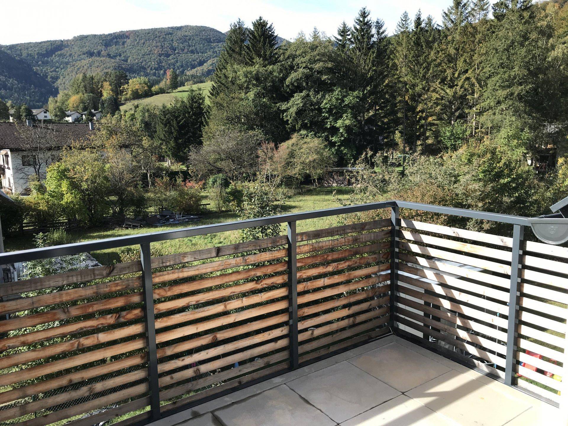 Sonniger Balkon mit Fliesenboden und Holzgeländer, mit Ausblick auf die grüne Hügellandschaft.