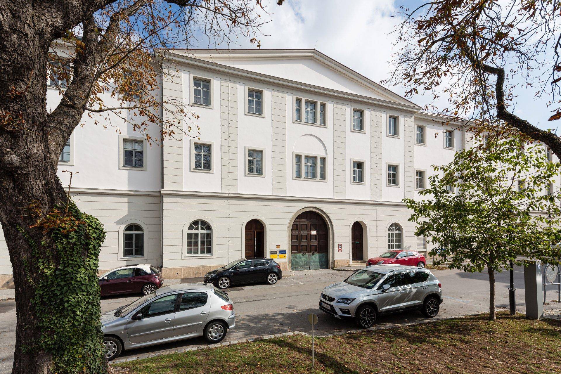 Klassische Fassade eines weißen Gebäudes mit geparkten Autos und Bäumen im Vordergrund.